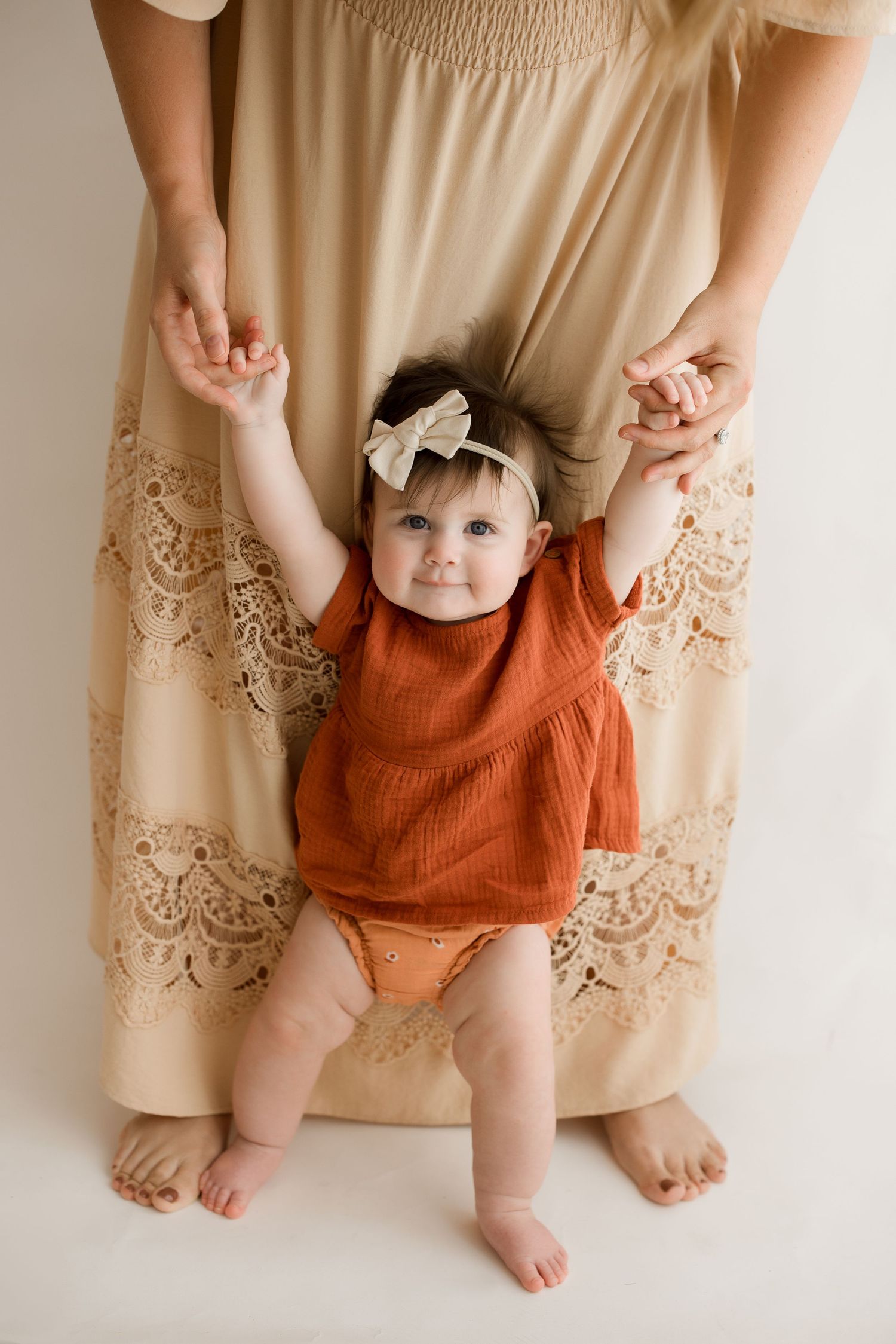 Baby in orange romper plays near parent wearing beige lace dress against neutral background.