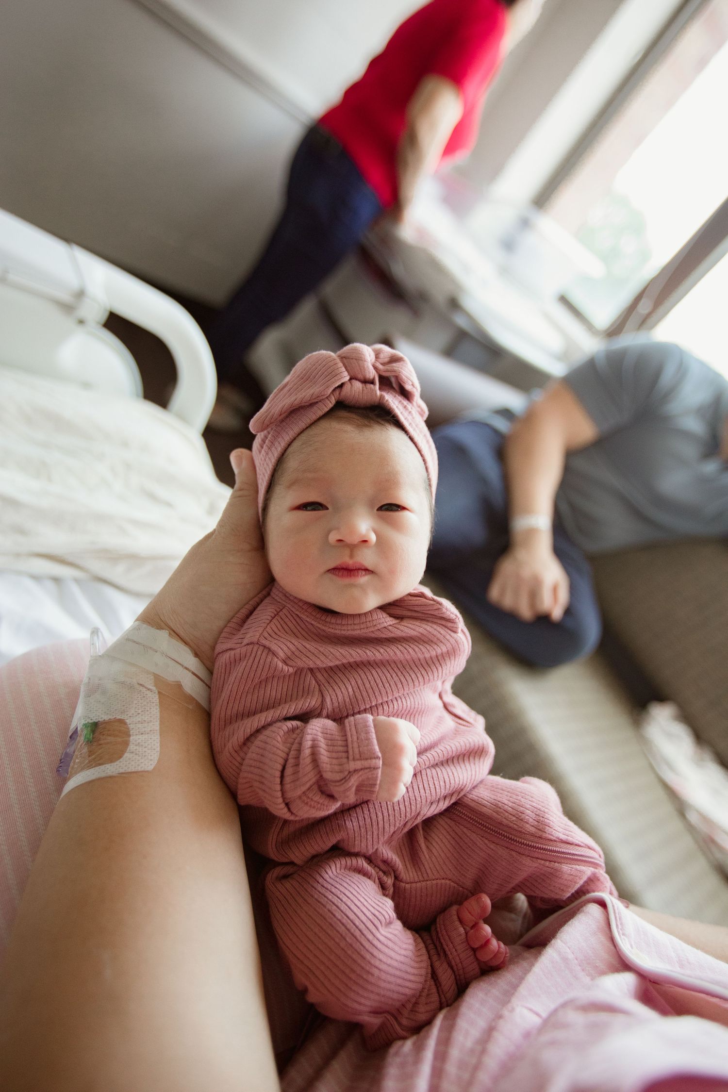 Baby wearing a pink ribbed outfit and headband rests on someone's legs.