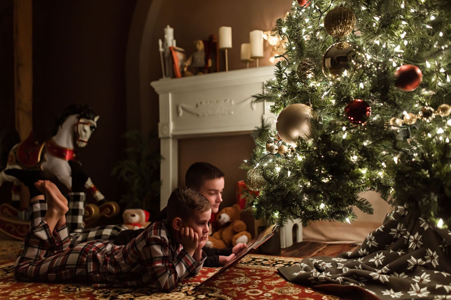 A Christmas tree illuminates the corner of a room with sparkling lights and ornaments near a classic white fireplace.