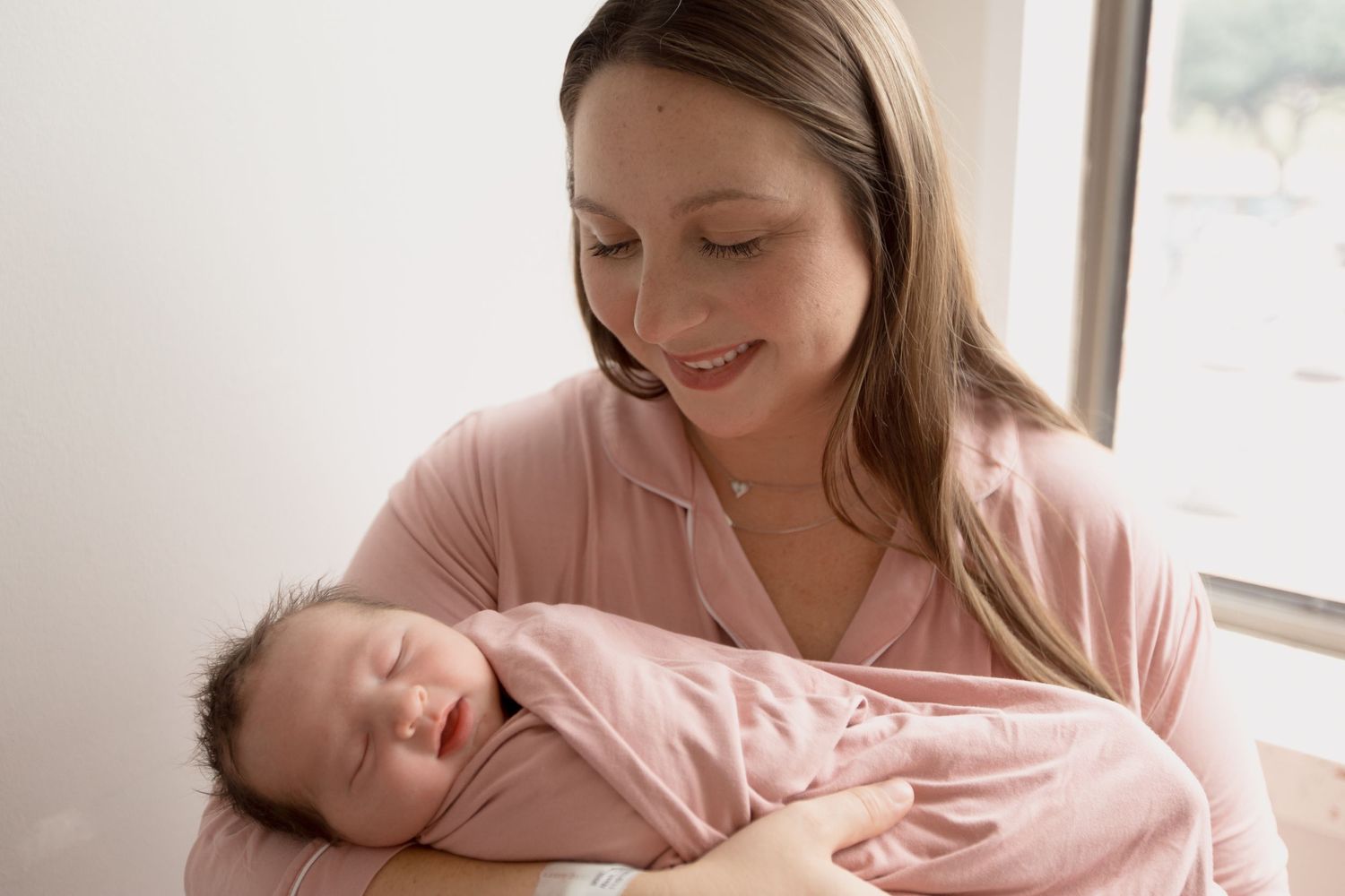 A mother in a pink sweater holds and gazes lovingly at her sleeping baby near a window.