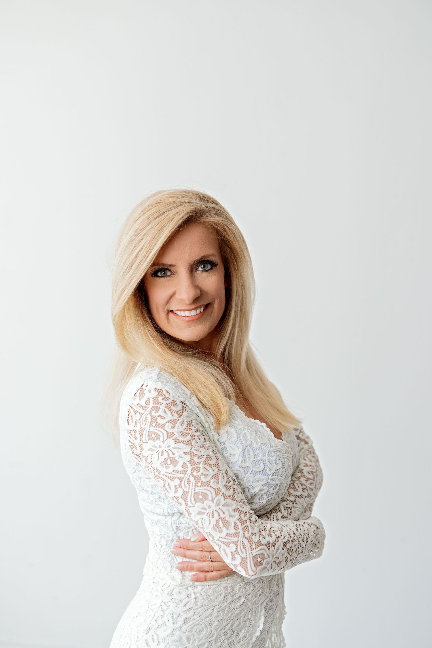 A blonde woman in a white lace long sleeve dress poses against a light background.