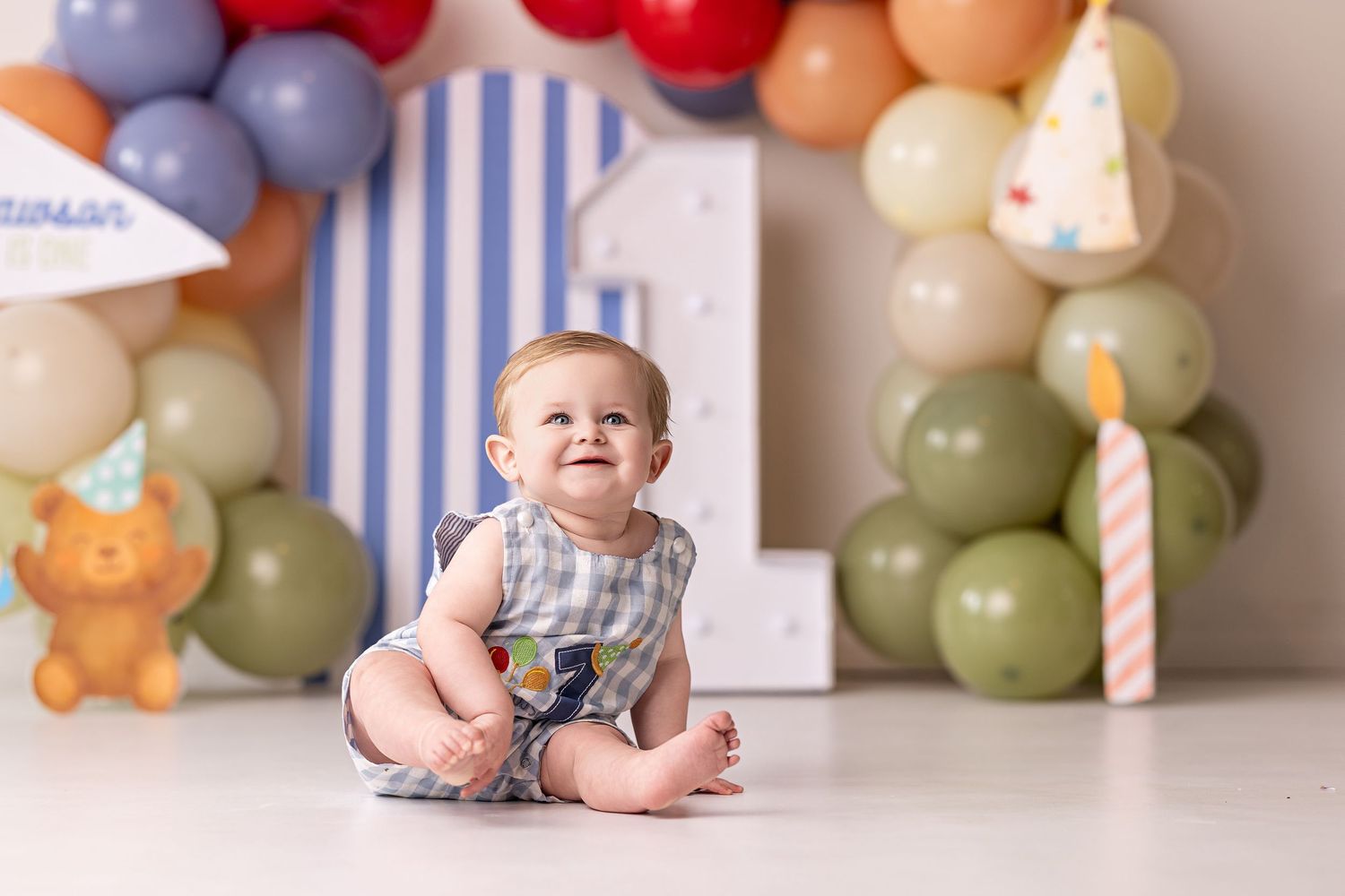 A baby's cake smash photo session with colorful balloon decorations and a blue and white striped backdrop.