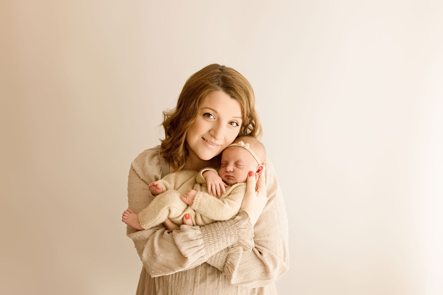 Mother in beige sweater holding and cuddling newborn baby against neutral backdrop.