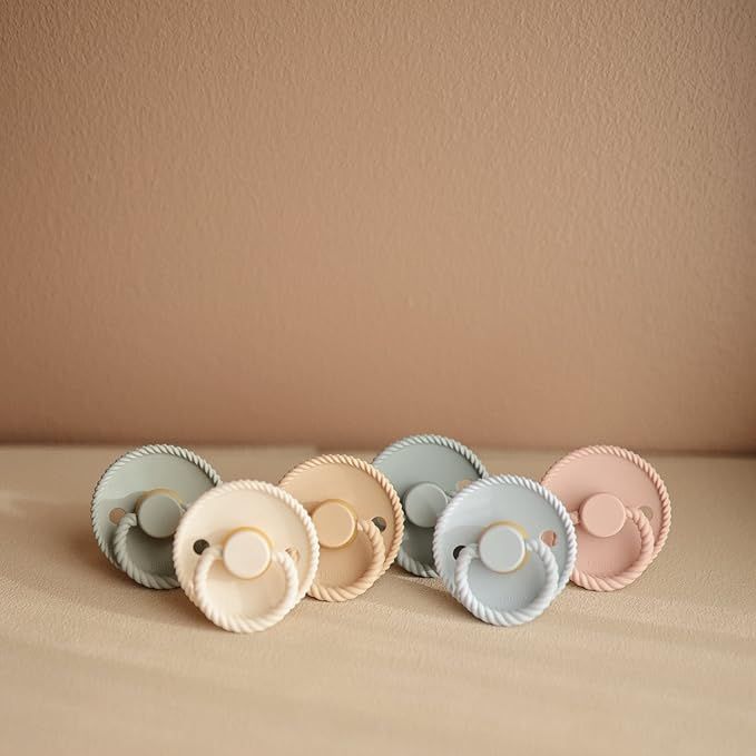 Modern pastel silicone baby pacifiers arranged in soft pink, beige and gray colors against a neutral background.