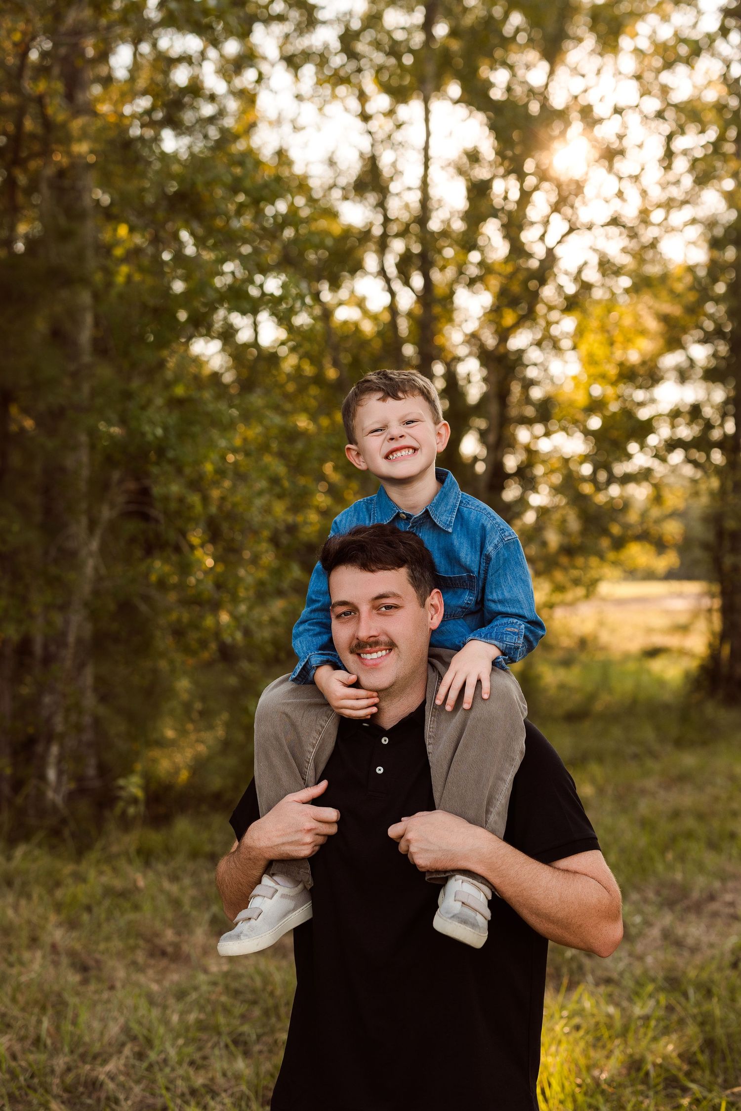 Smiling young boy in a blue denim shirt sits on the shoulders of a mustached man in black in a sunlit grassy field with trees.