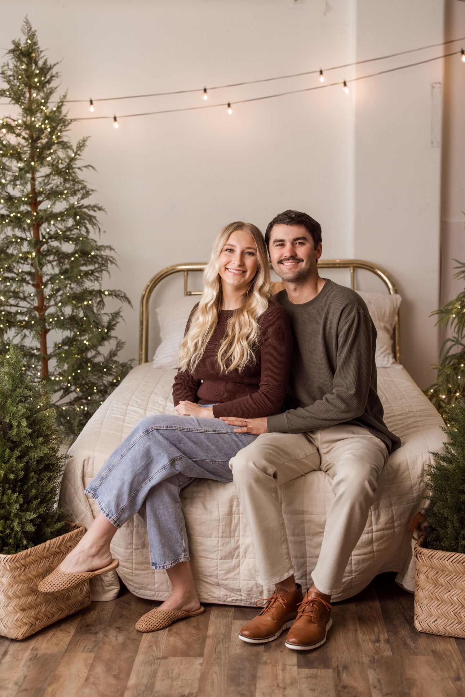 Cozy holiday scene with couple sitting on white armchair between Christmas trees and string lights on wall.