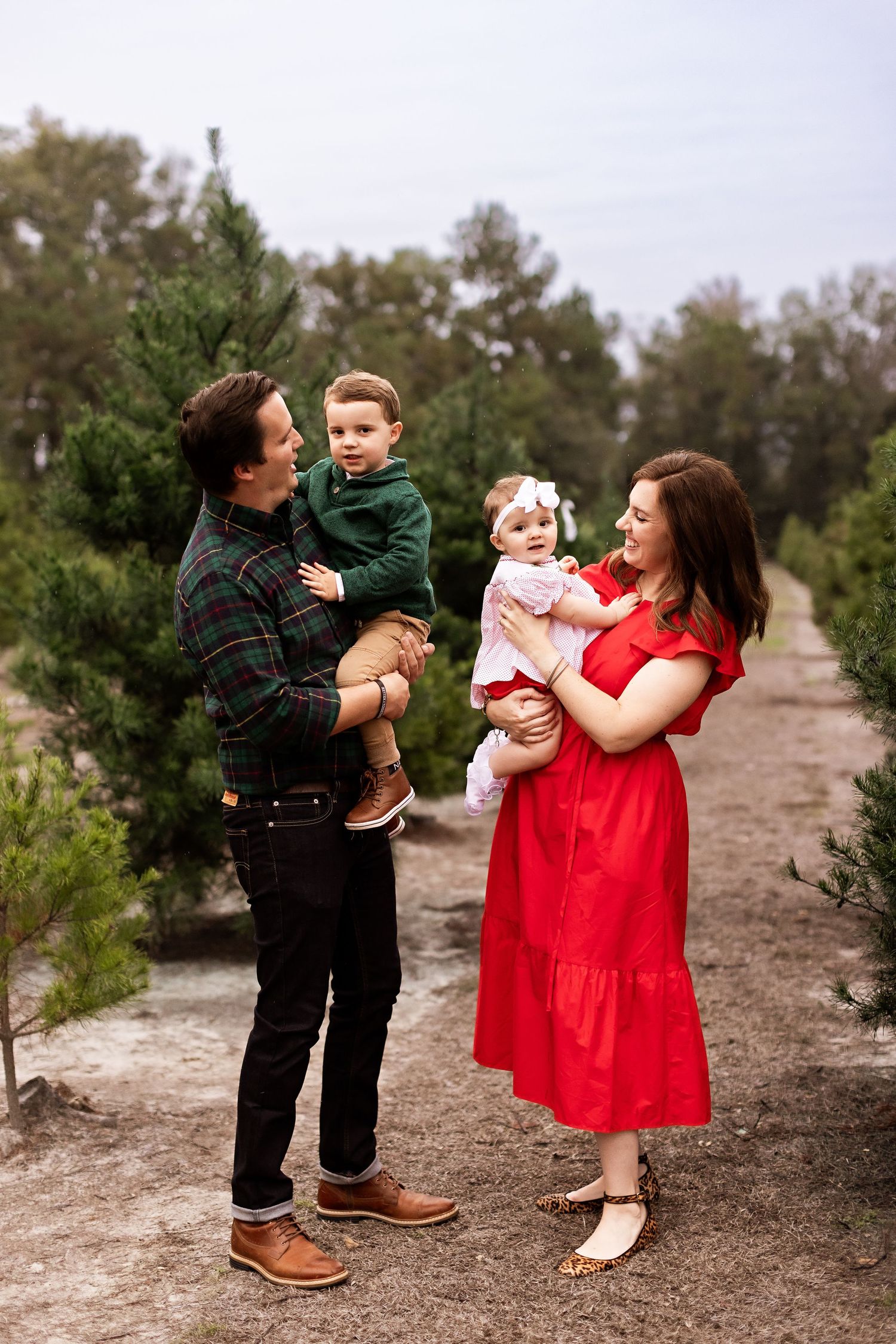 A family of four poses together at a Christmas tree farm with evergreen trees in the background.