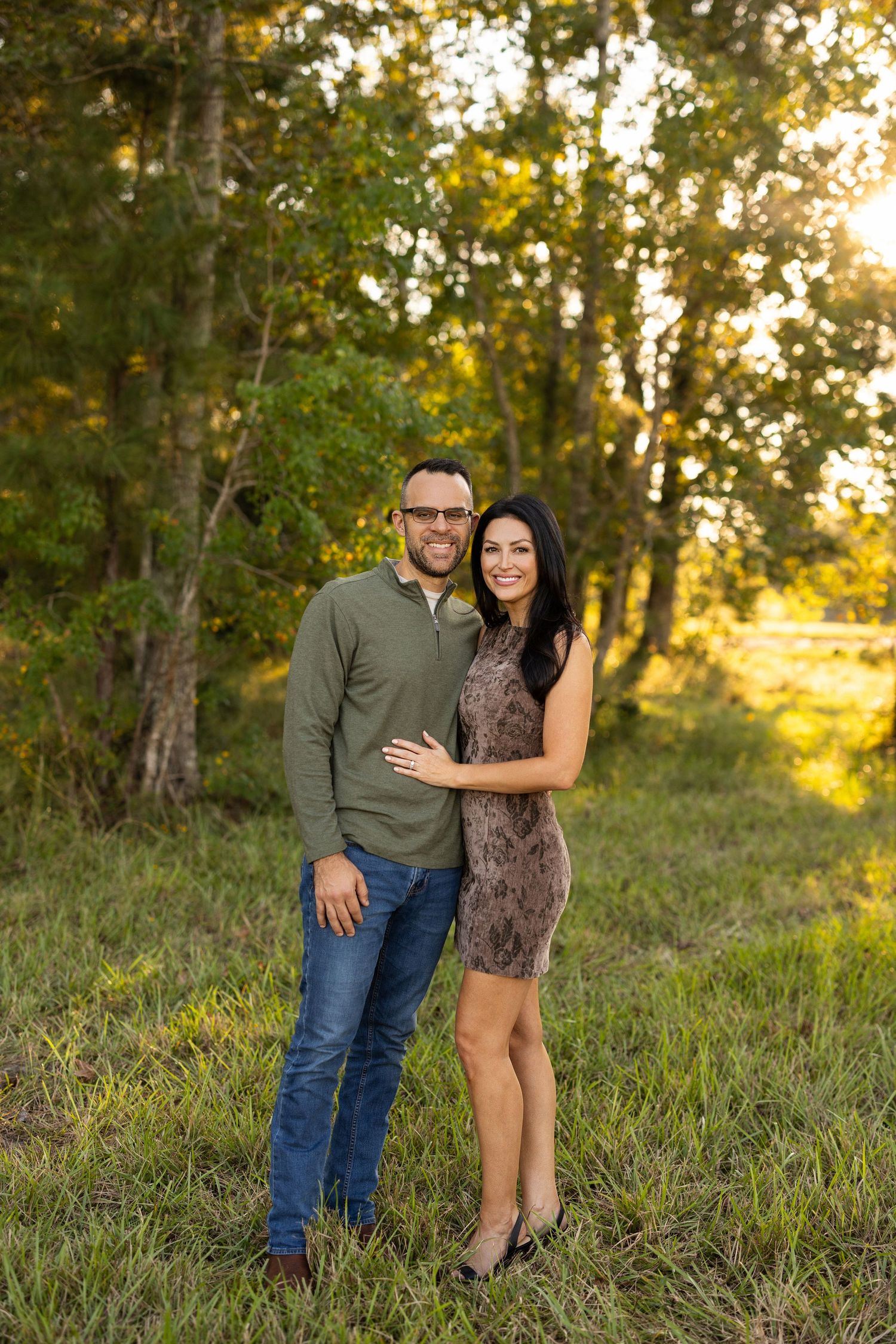 A couple stands together in a wooded area during golden hour.