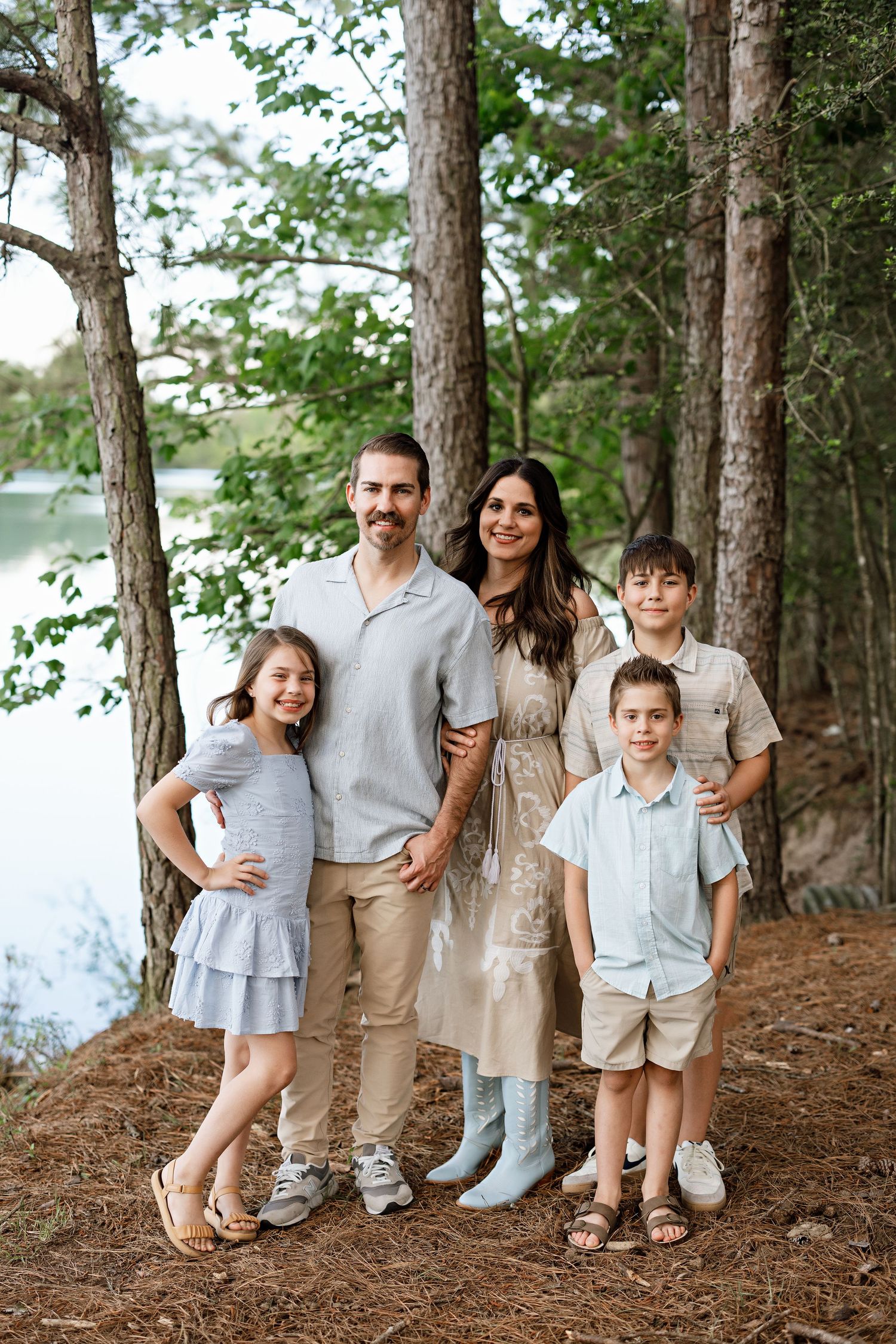 A family poses together on a wooded trail by a serene lake wearing coordinated neutral outfits.
