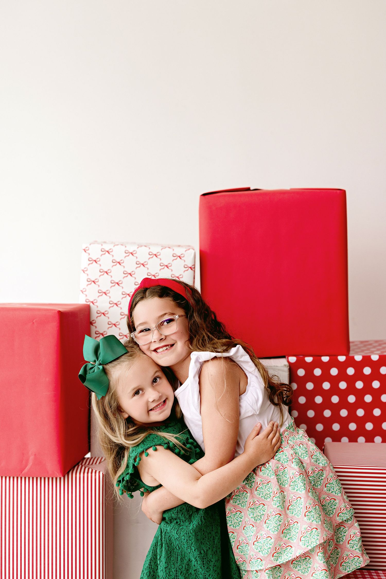Two children embrace while posing in front of red and white wrapped Christmas presents and gift boxes.