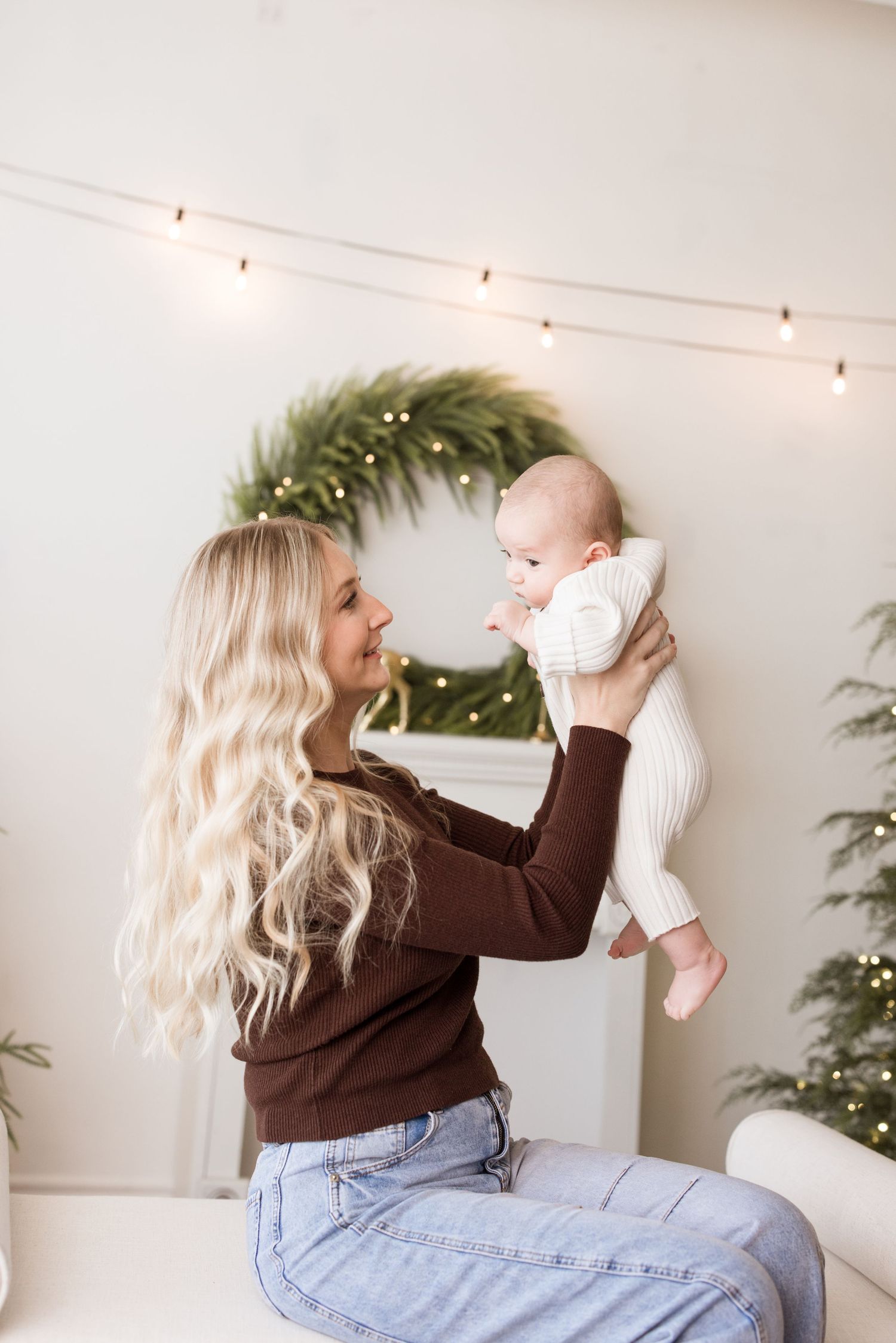 Woman in brown sweater and jeans lifts and plays with baby in bright interior setting with string lights.