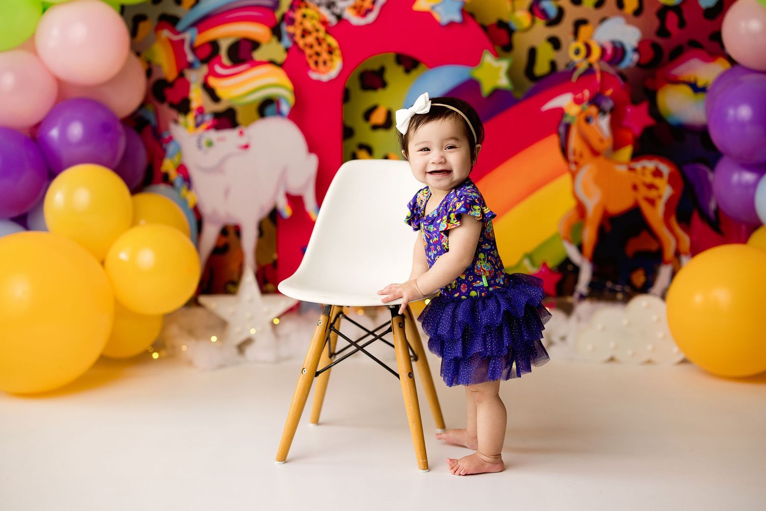 Colorful party scene with balloons, unicorn decorations, and a white eames-style chair against a rainbow backdrop.