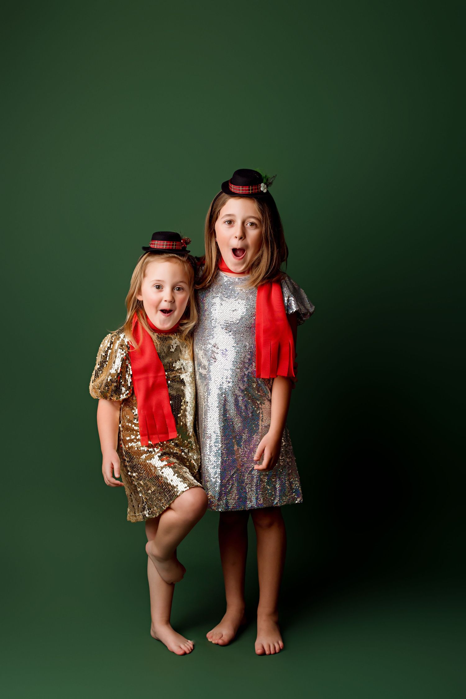 Two children in festive sequin dresses and red scarves pose excitedly against green backdrop.