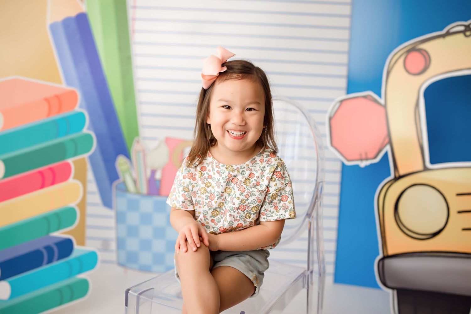 Child sits smiling on chair against colorful school-themed backdrop with pencil illustrations.