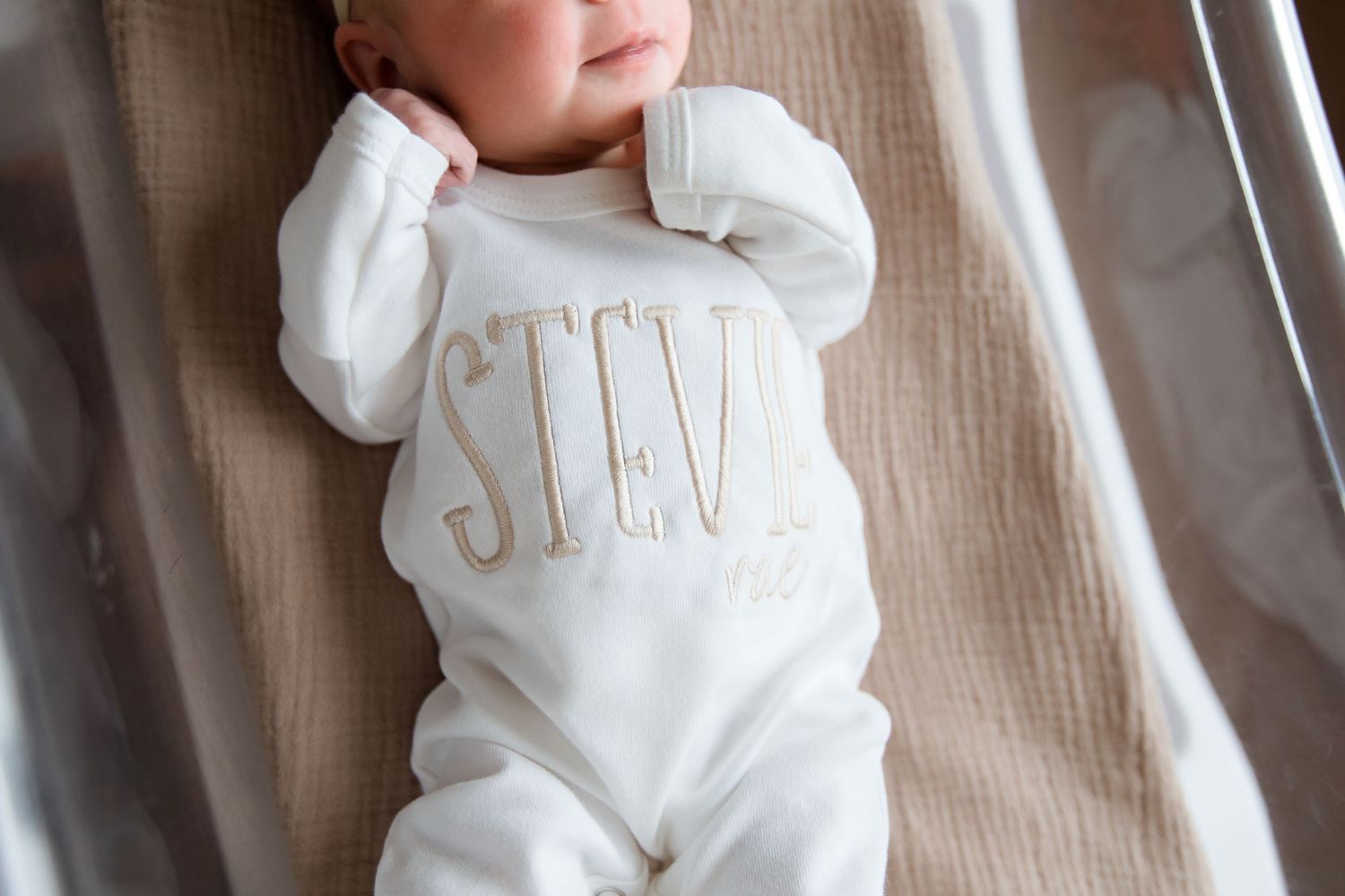 Newborn baby wearing white onesie with embroidered text resting on beige fabric during Fresh 48 session in Beaumont hospital.