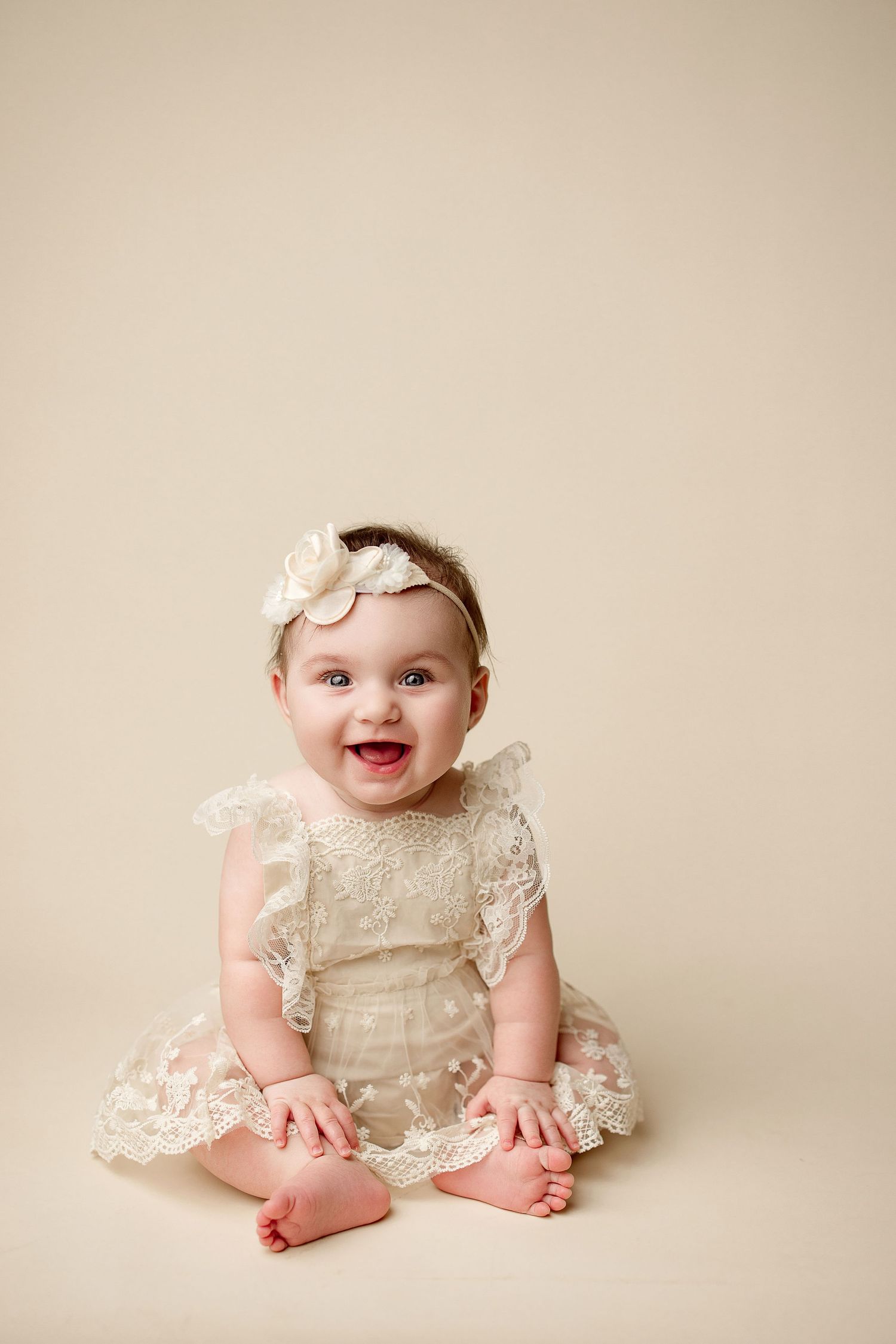 A baby wearing a vintage white lace dress and headband sits on a beige backdrop in a sweet portrait series.