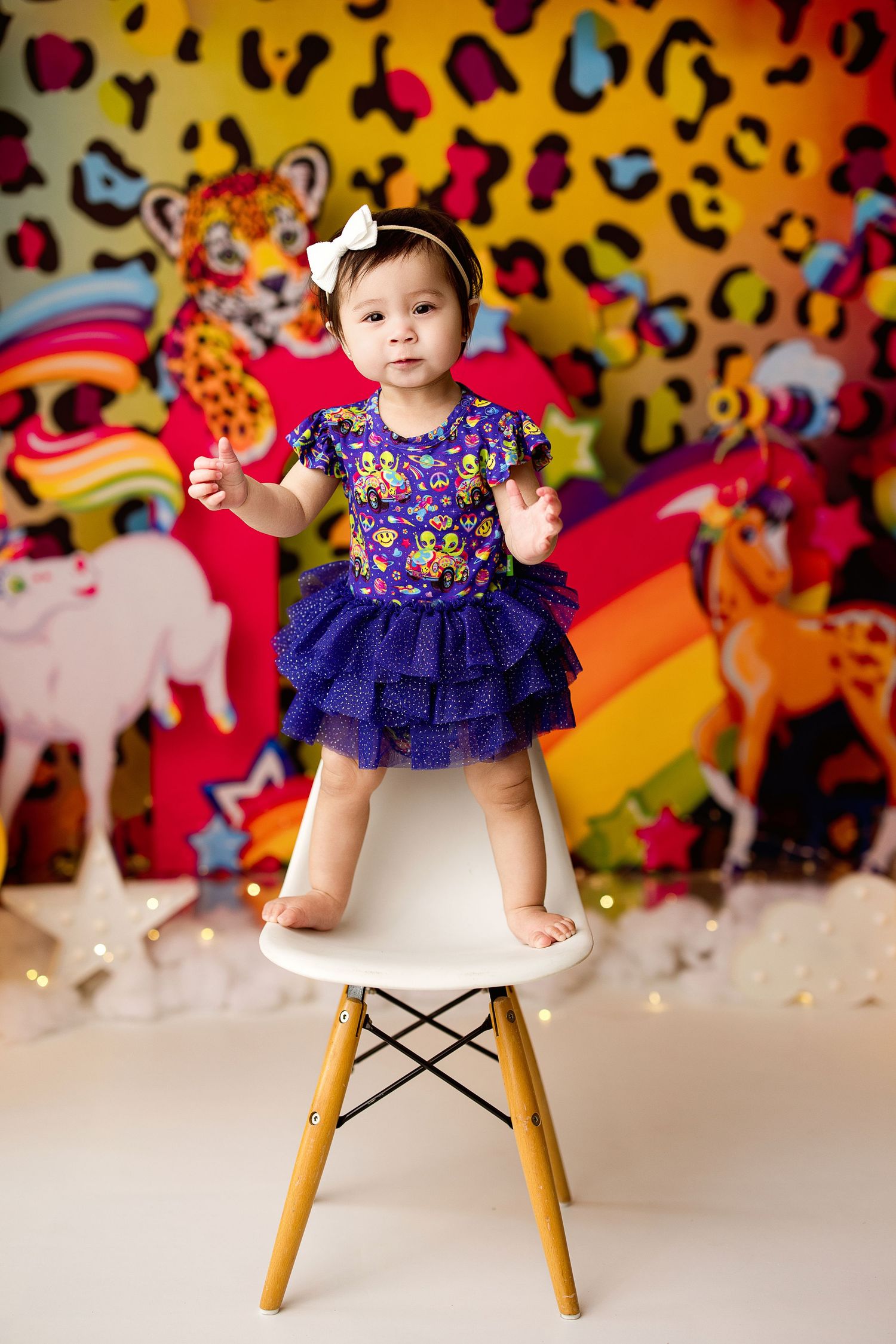 Small child stands on white chair against colorful leopard print backdrop with rainbow stripes and animal decorations.