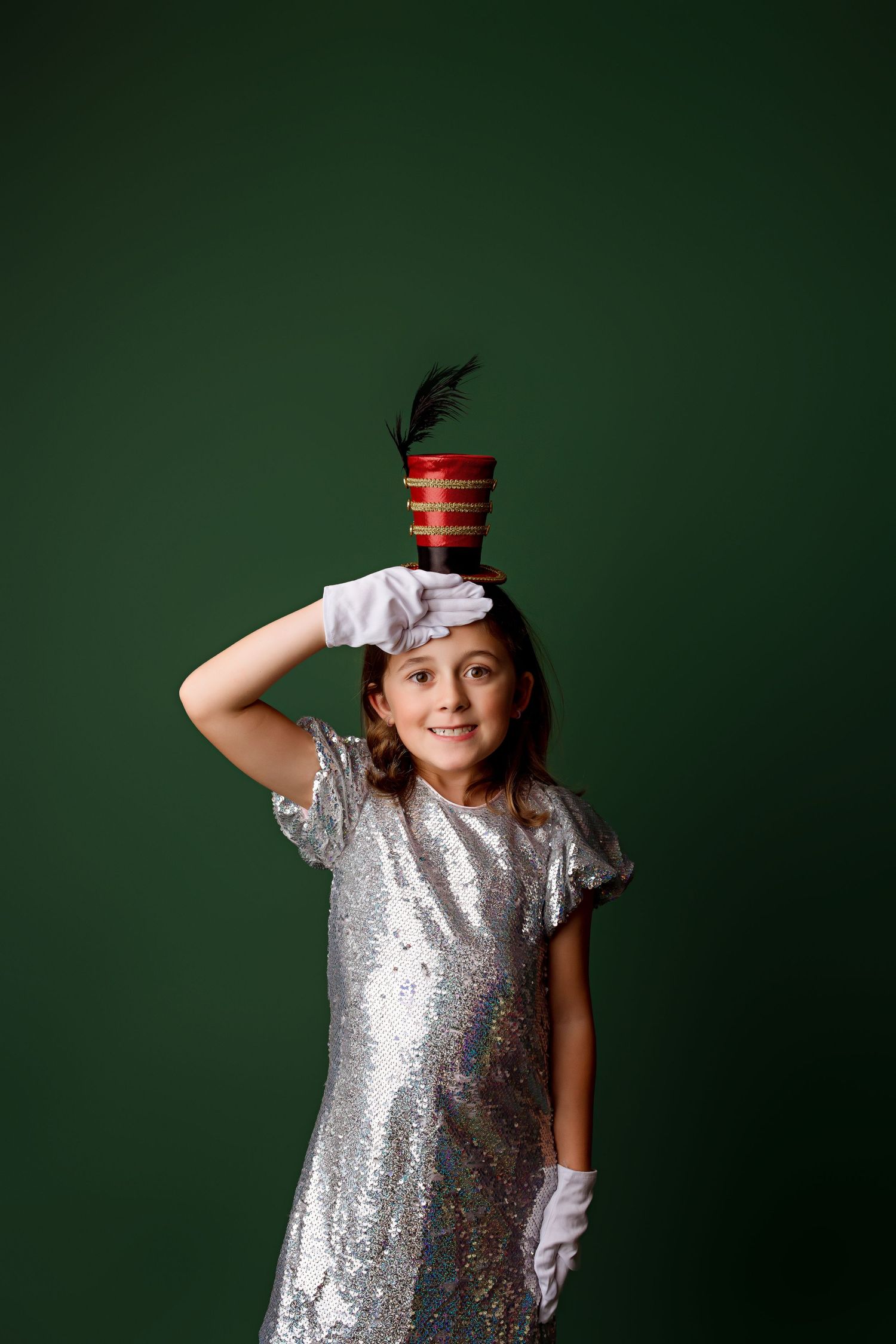 Child in sparkly silver dress poses with toy soldier hat against green background.