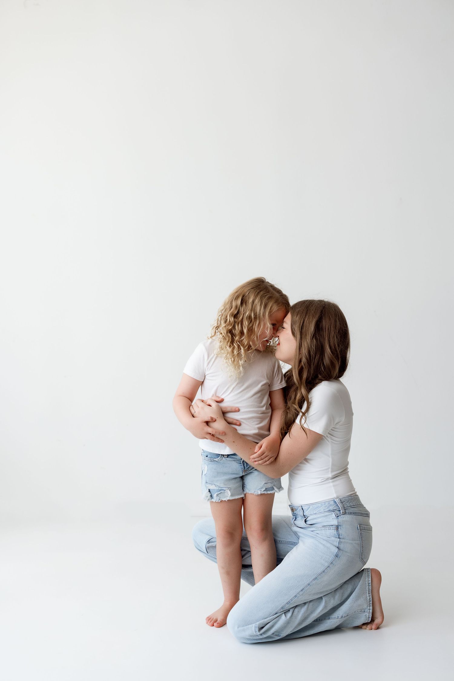Parent and young child share an intimate moment of affection against a white backdrop in casual blue jeans and white tops.