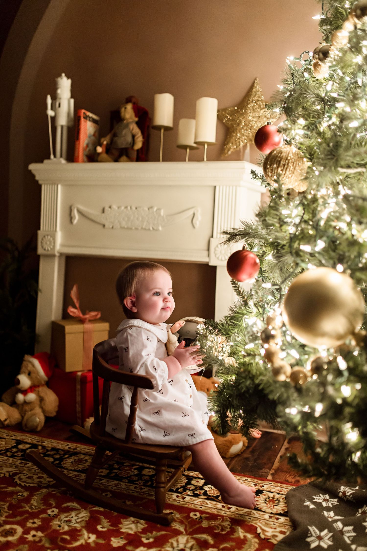 A richly decorated Christmas tree glows warmly next to a white fireplace mantel in a cozy living room setting.