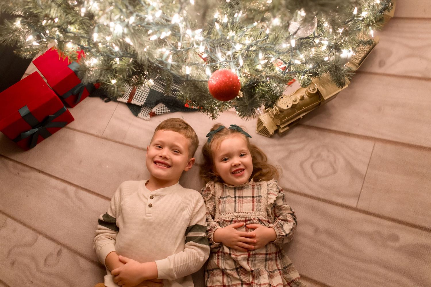 Children sit together and laugh while posing in front of a decorated Christmas tree with twinkling lights.