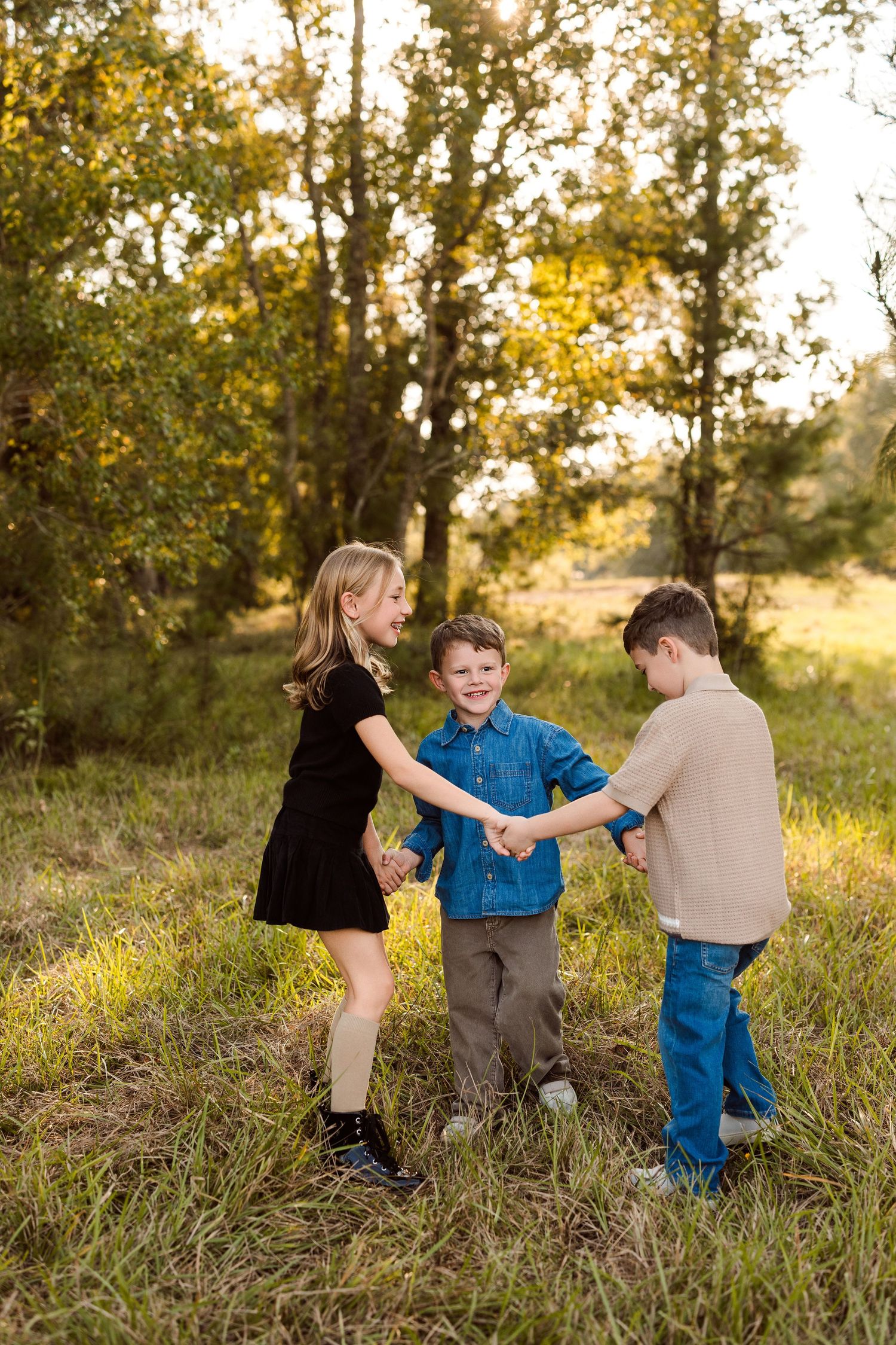 Three children hold hands in a circle in a sunny grassy field with trees, smiling and playing during a golden hour family photo session.