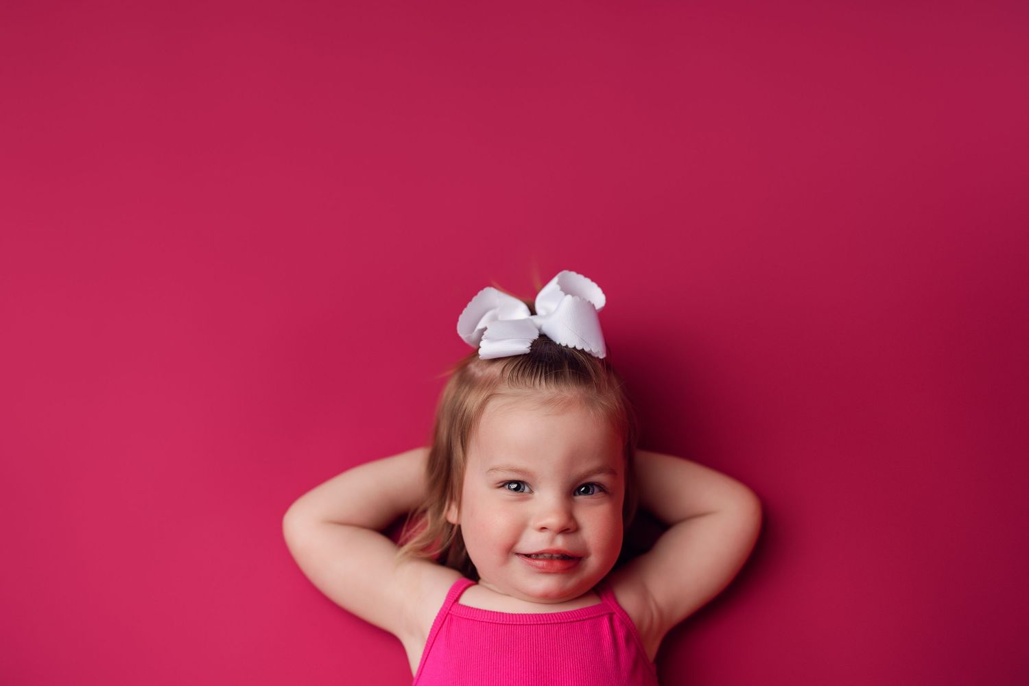 Smiling child with white bow on head against pink background.