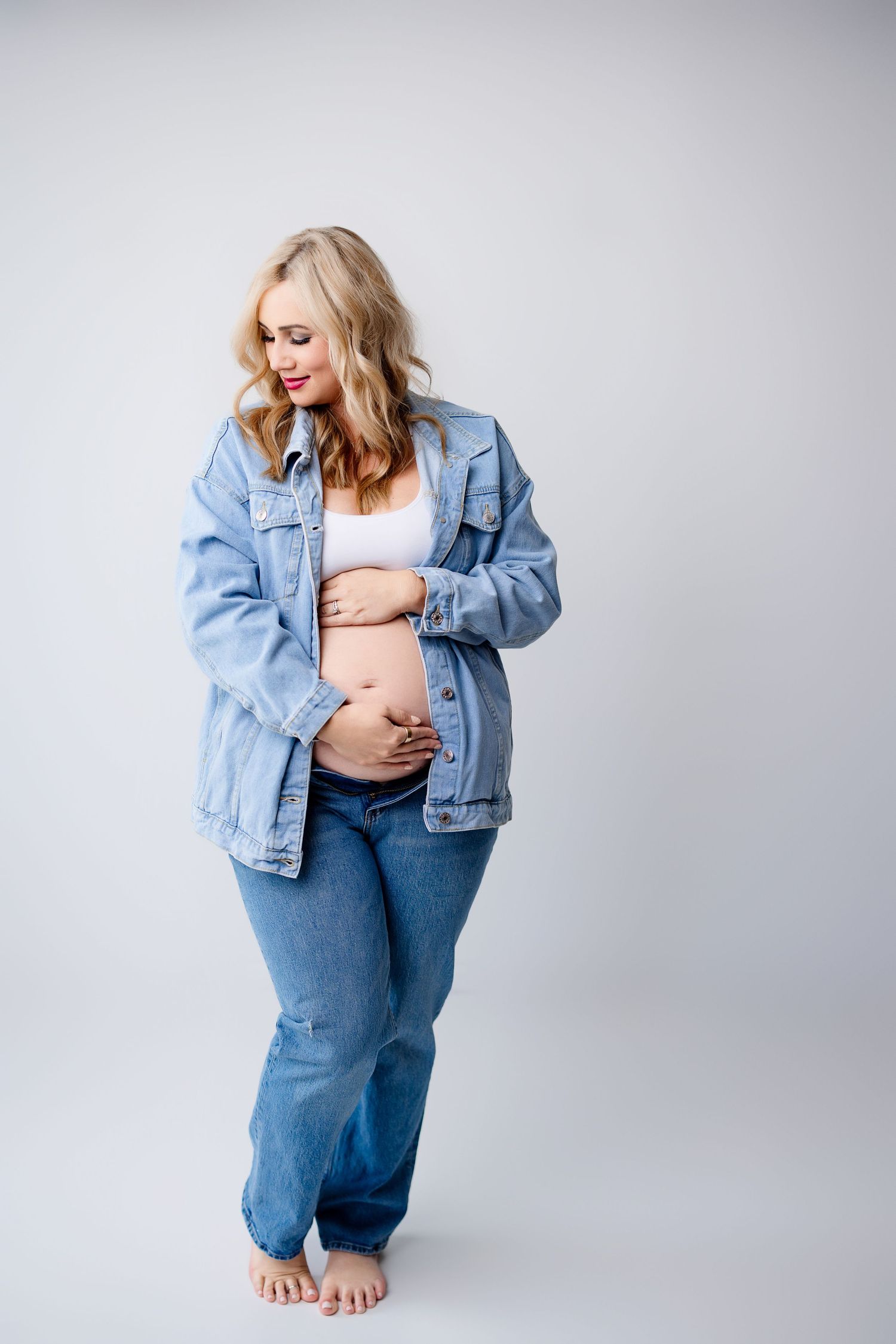 An expectant mother in denim jacket and jeans poses against a light gray studio background with Bree Elle Photography in Port Neches, TX