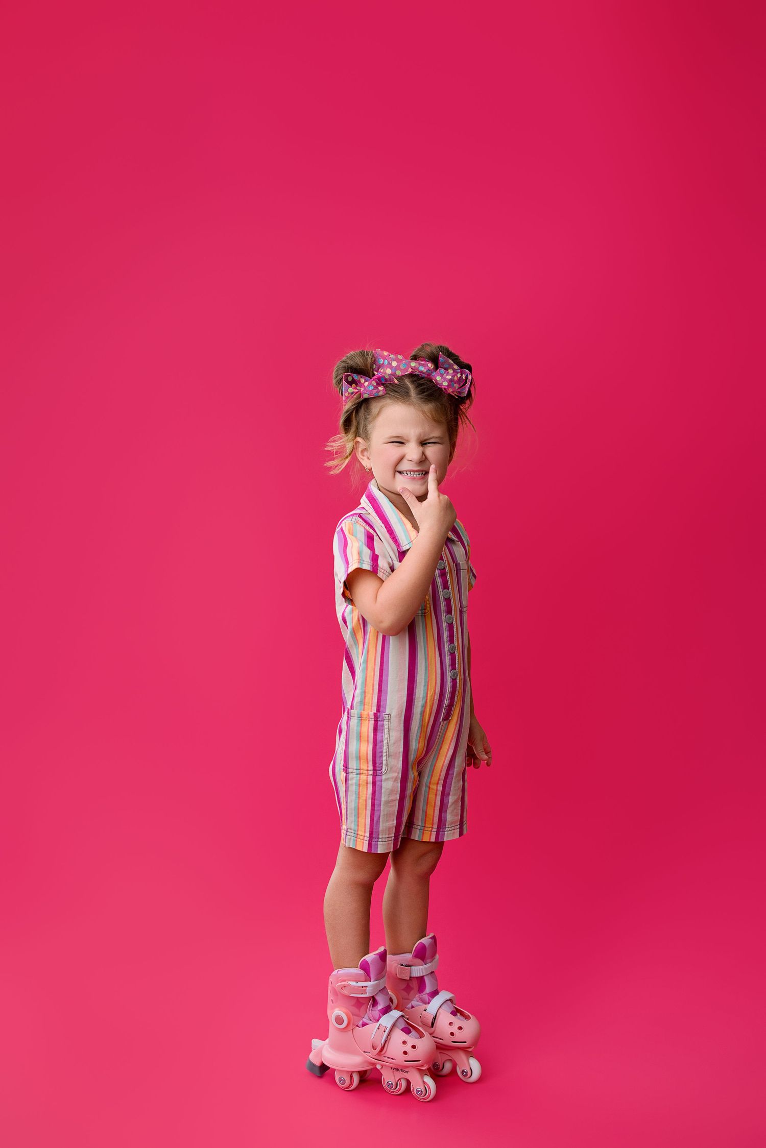 A child in colorful striped clothing and roller skates poses playfully against a bright pink studio background.