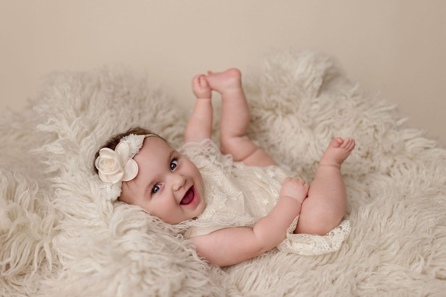 A joyful baby laughs while lying on a soft white fur rug during a newborn photo session.
