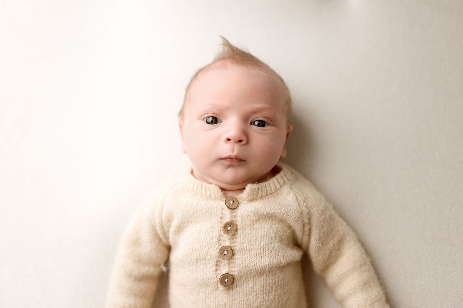 A baby wearing a cozy cream-colored cardigan sweater with wooden buttons lies on a white background.