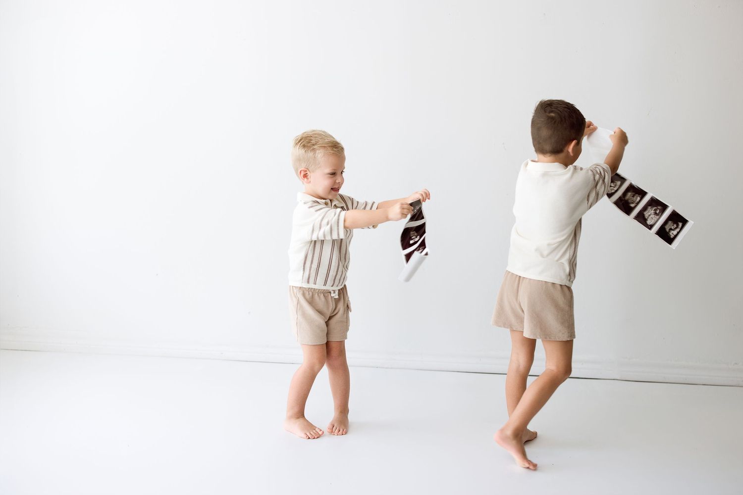 Two small children wearing beige outfits playing while holding toy cameras in a white minimalist setting.