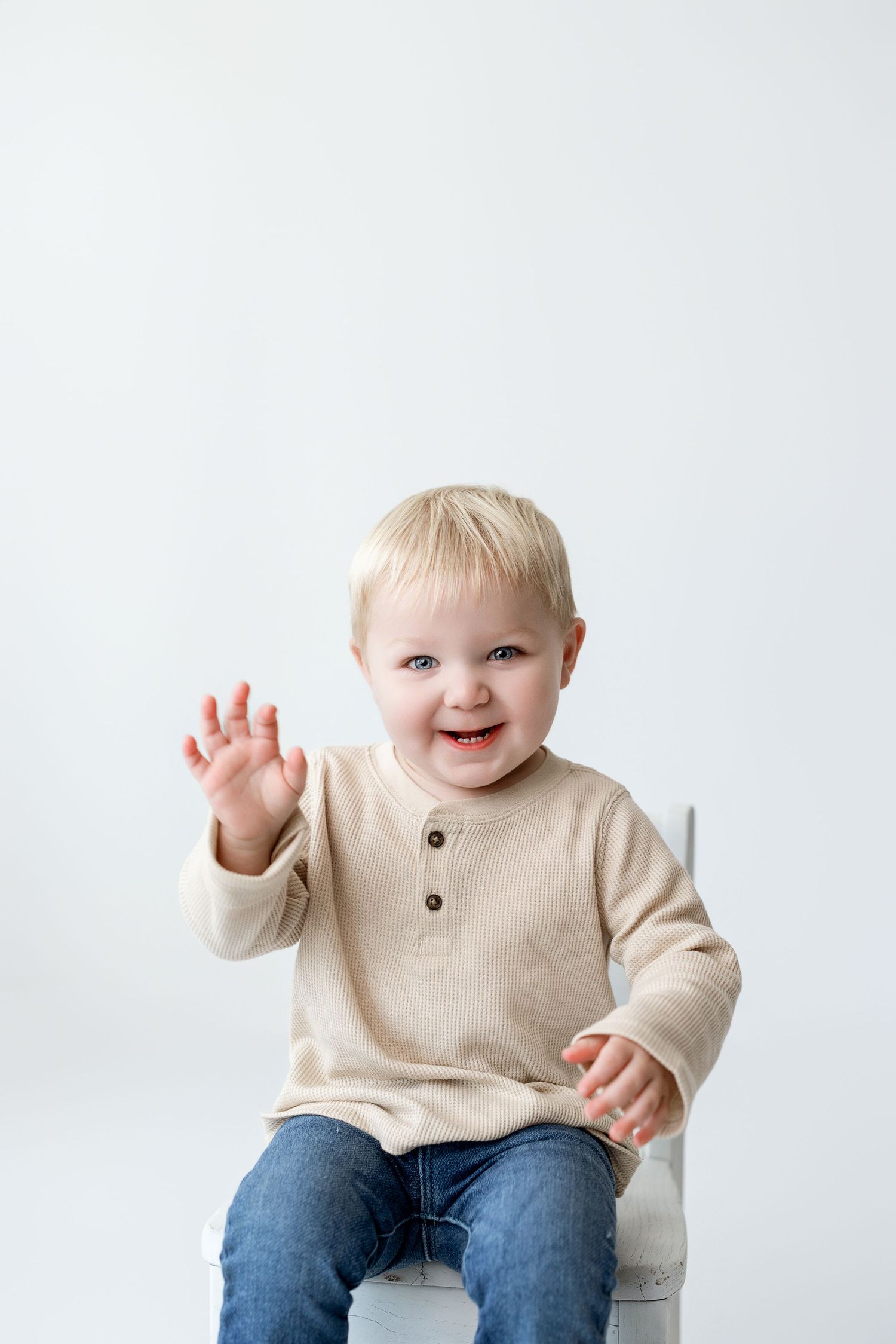 Happy toddler in beige sweater and jeans waves during photo series against white background.