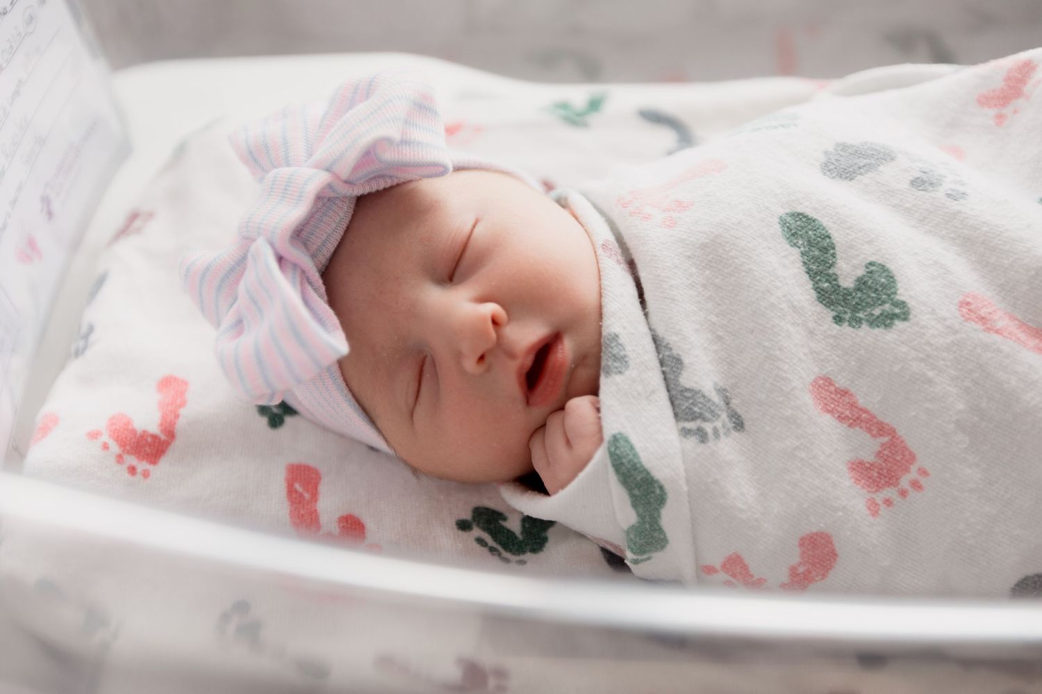 Sleeping newborn swaddled in white blanket with pink and green dinosaur prints and wearing light pink bow headband.