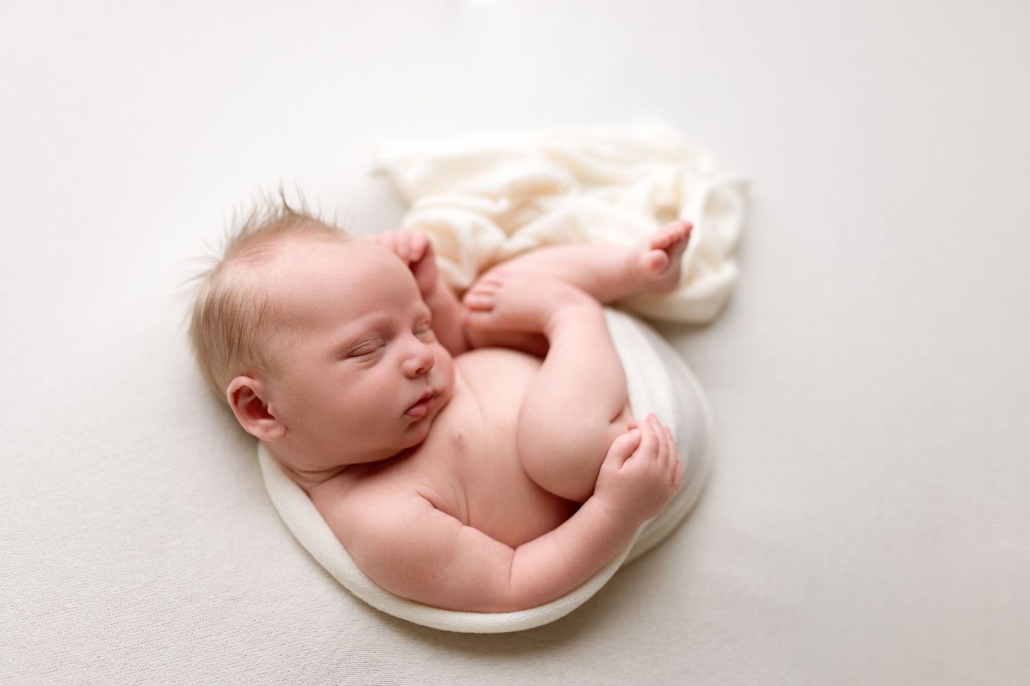 A sleeping newborn baby lies peacefully on soft white fabric in a bright, minimalist setting.