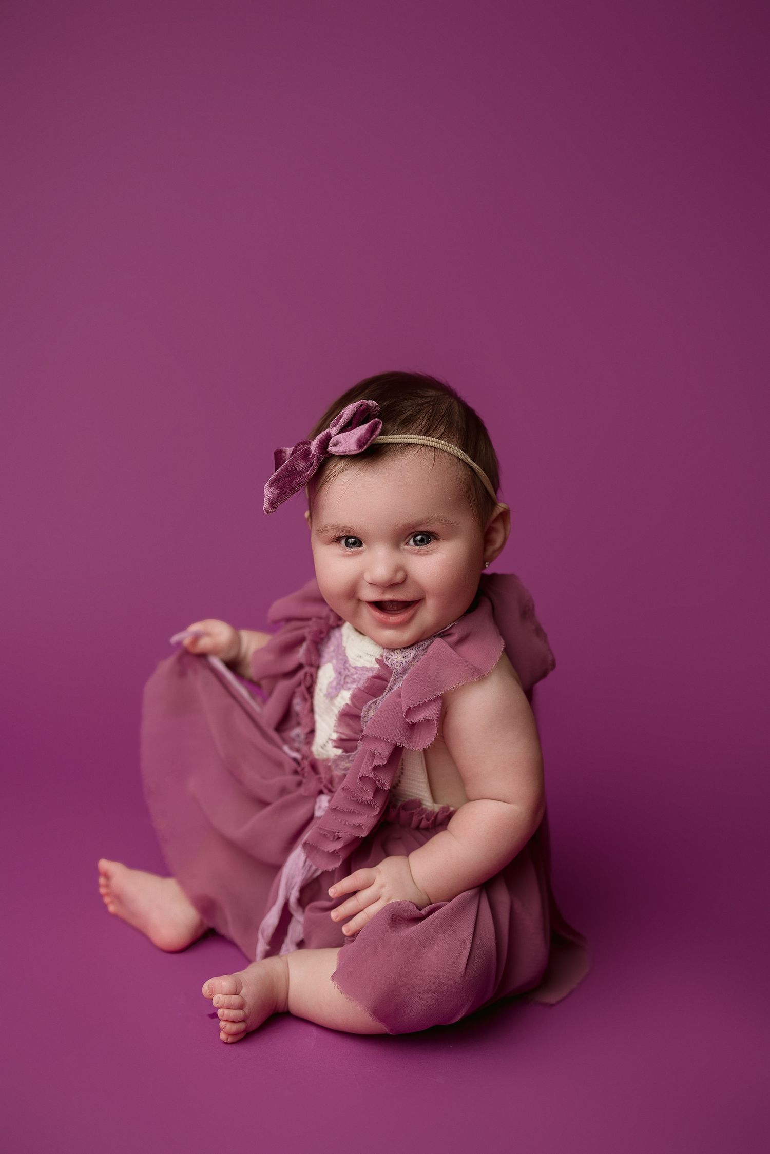 Baby in dusty rose colored dress and matching headband sits on pink backdrop smiling at camera.