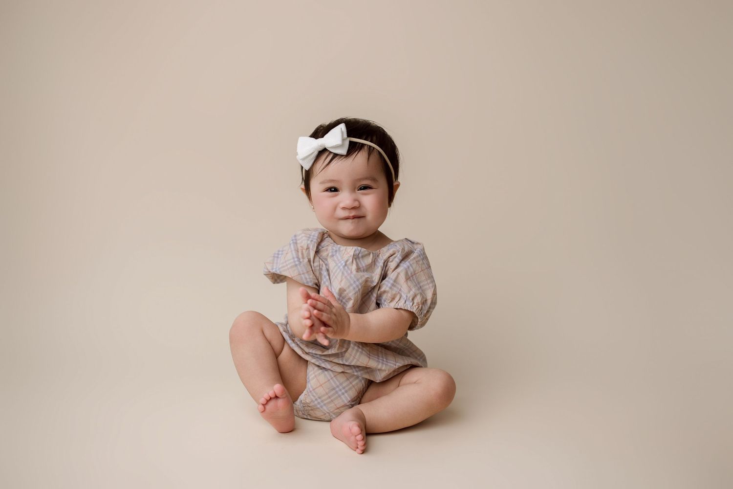 Adorable baby in a beige romper sits on light beige backdrop looking softly downward at viewer.