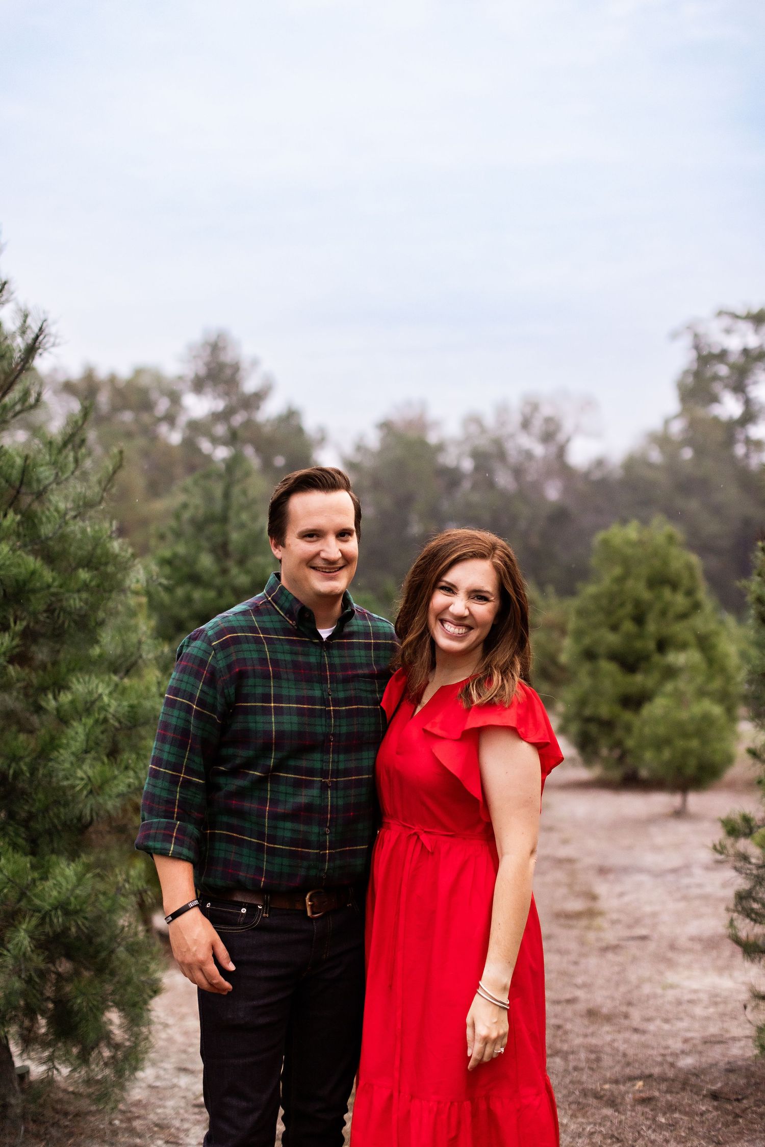 A couple poses together outdoors at a tree farm, with her wearing a red dress and him in a plaid shirt.