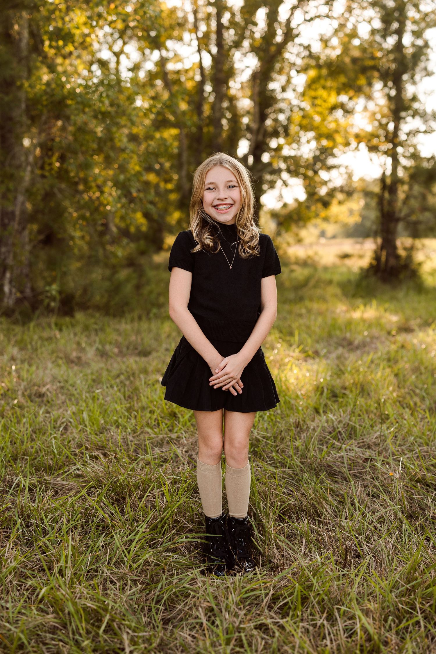 Smiling girl in a black short-sleeve top and skirt with tall socks and boots stands in a sunlit grassy field with trees behind her.
