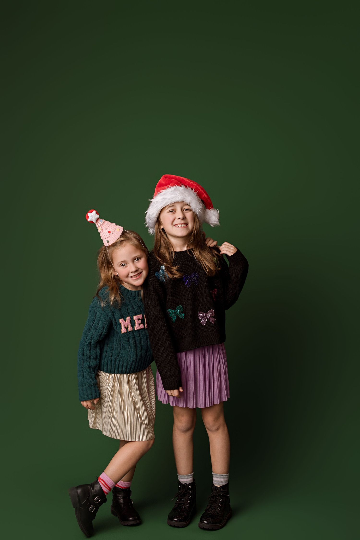 Two children in festive holiday outfits with Santa and party hats pose playfully against a dark green background.