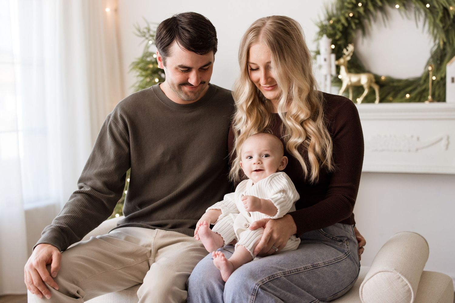 A cozy family portrait in front of a white fireplace with holiday wreath, featuring a baby in a white outfit.