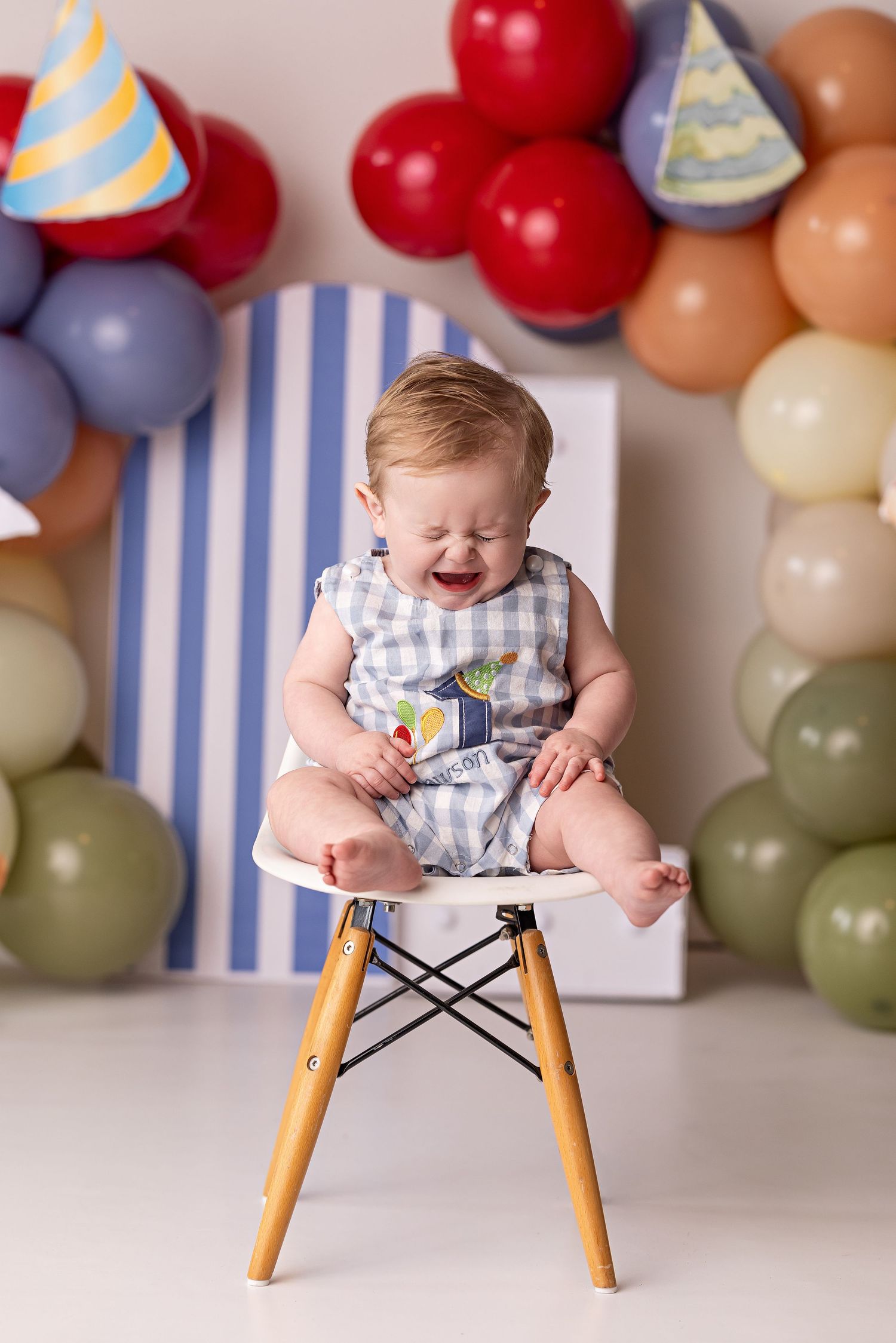 Baby in striped romper sitting on modern chair with colorful balloon arch and party decorations in background.