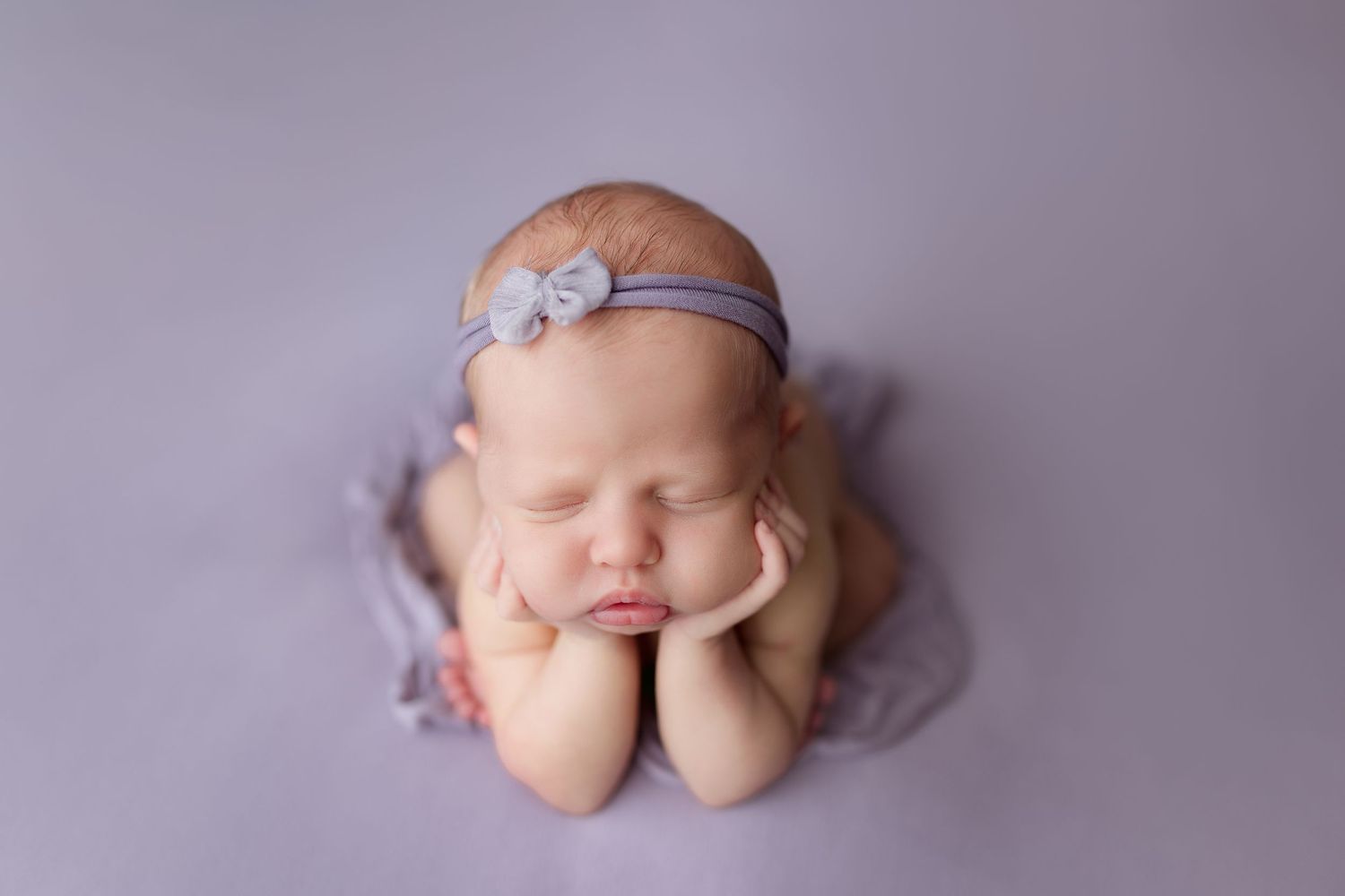 Sleeping newborn wearing delicate headband posed on purple backdrop.