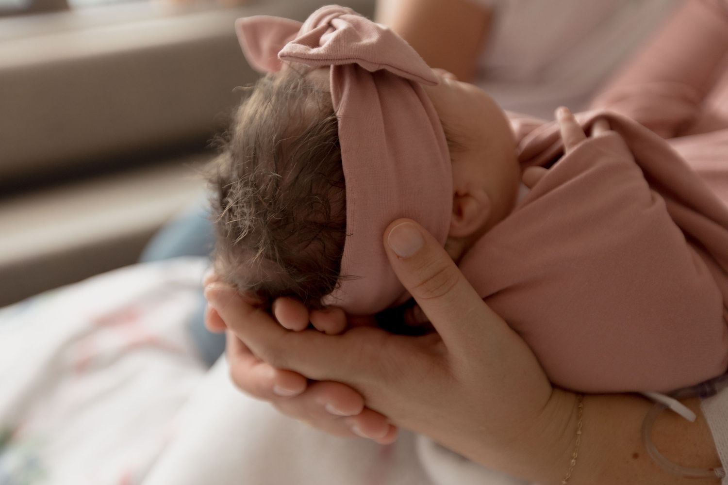 Mother tenderly cradling her sleeping newborn baby in soft pink tones by a sunlit window.