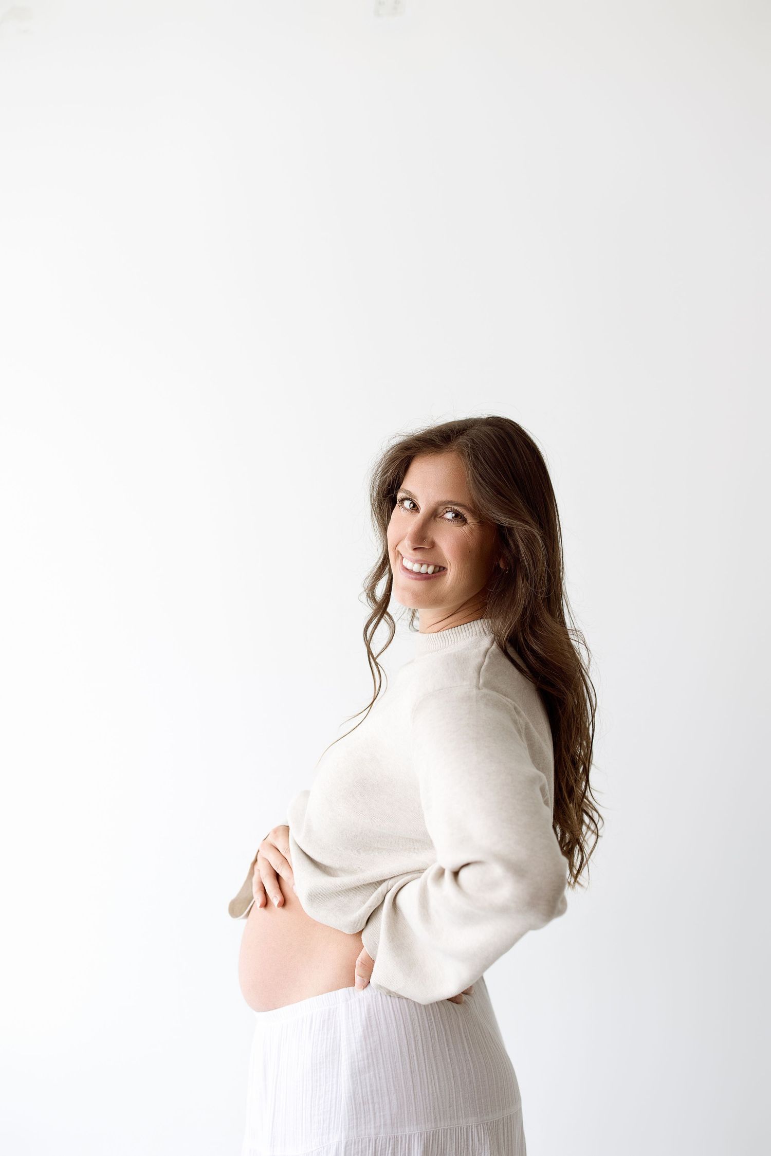 An expectant mother in a white sweater poses joyfully against a simple white background.