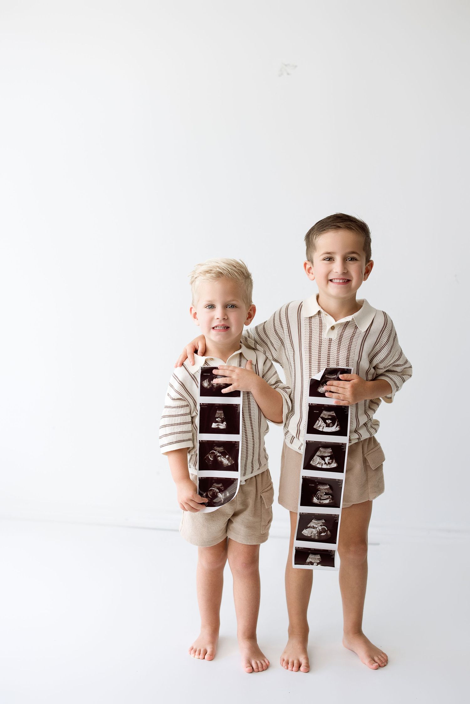 Two children in matching beige outfits hold up ultrasound photos while sharing excited smiles against a white backdrop.