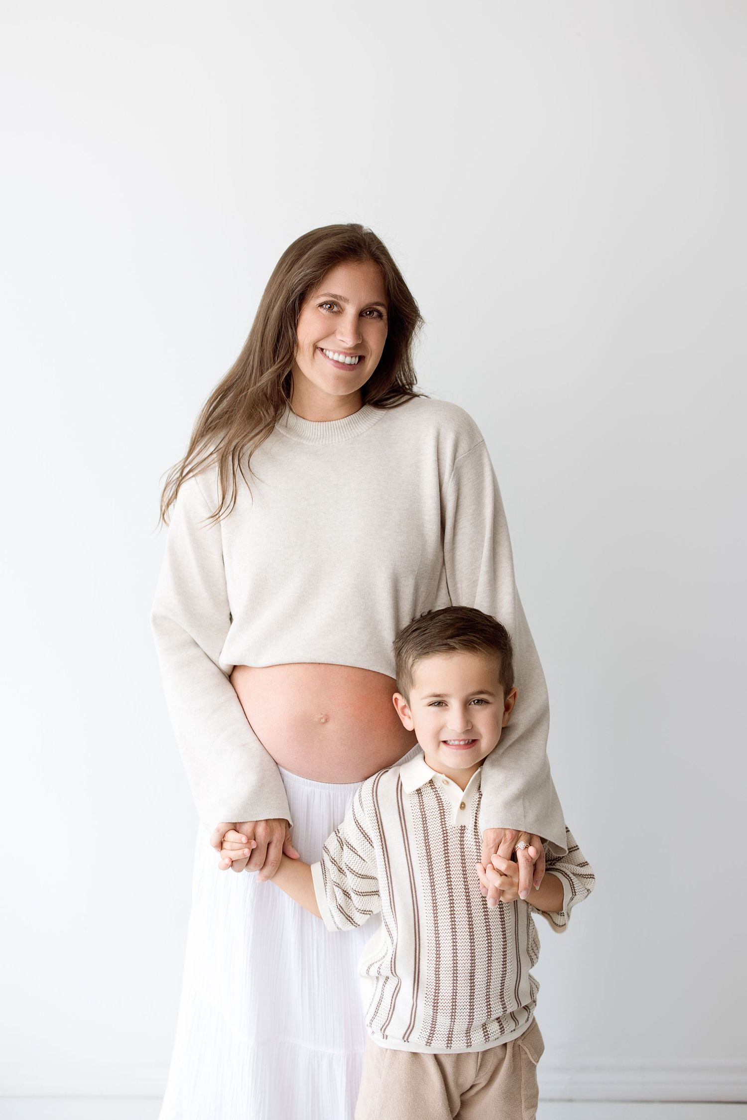 A pregnant woman in white clothing stands with a young child in a striped shirt against a white backdrop.