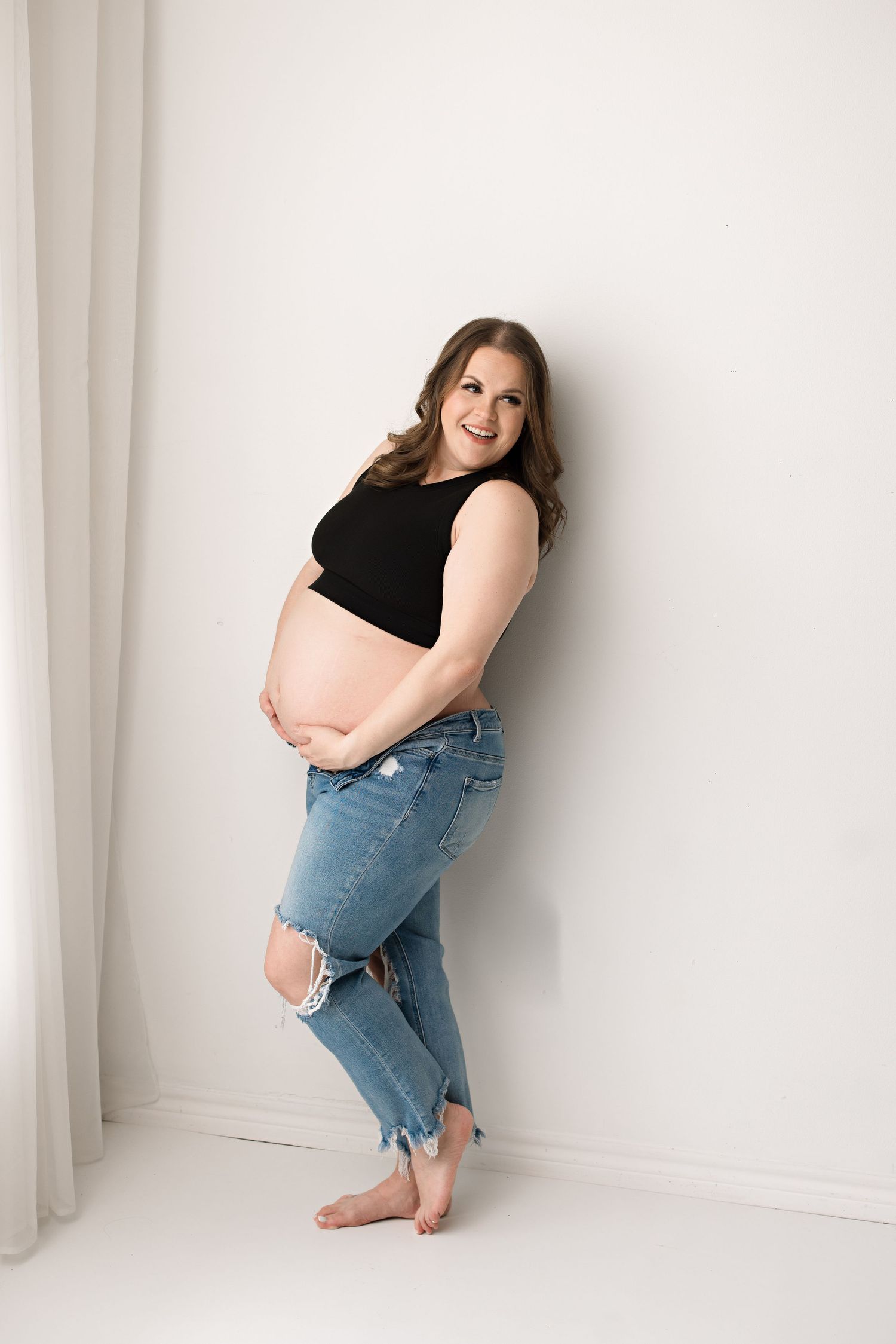 Casual maternity portrait in black top and distressed jeans against white backdrop with Bree Elle Photography in Beaumont, TX.