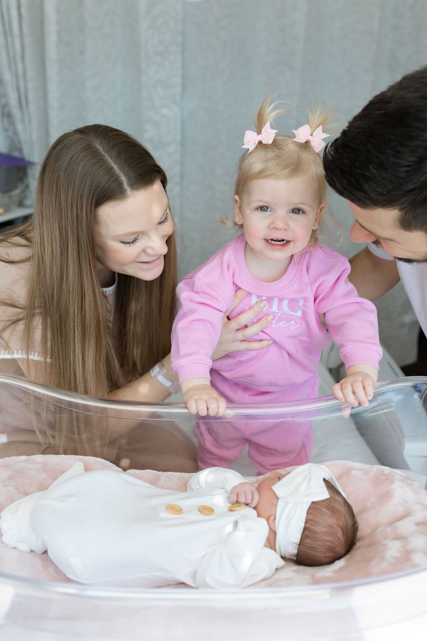 A family gathers around a newborn baby lying on pink bedding while a toddler in pink pajamas looks on with excitement during Fresh 48.