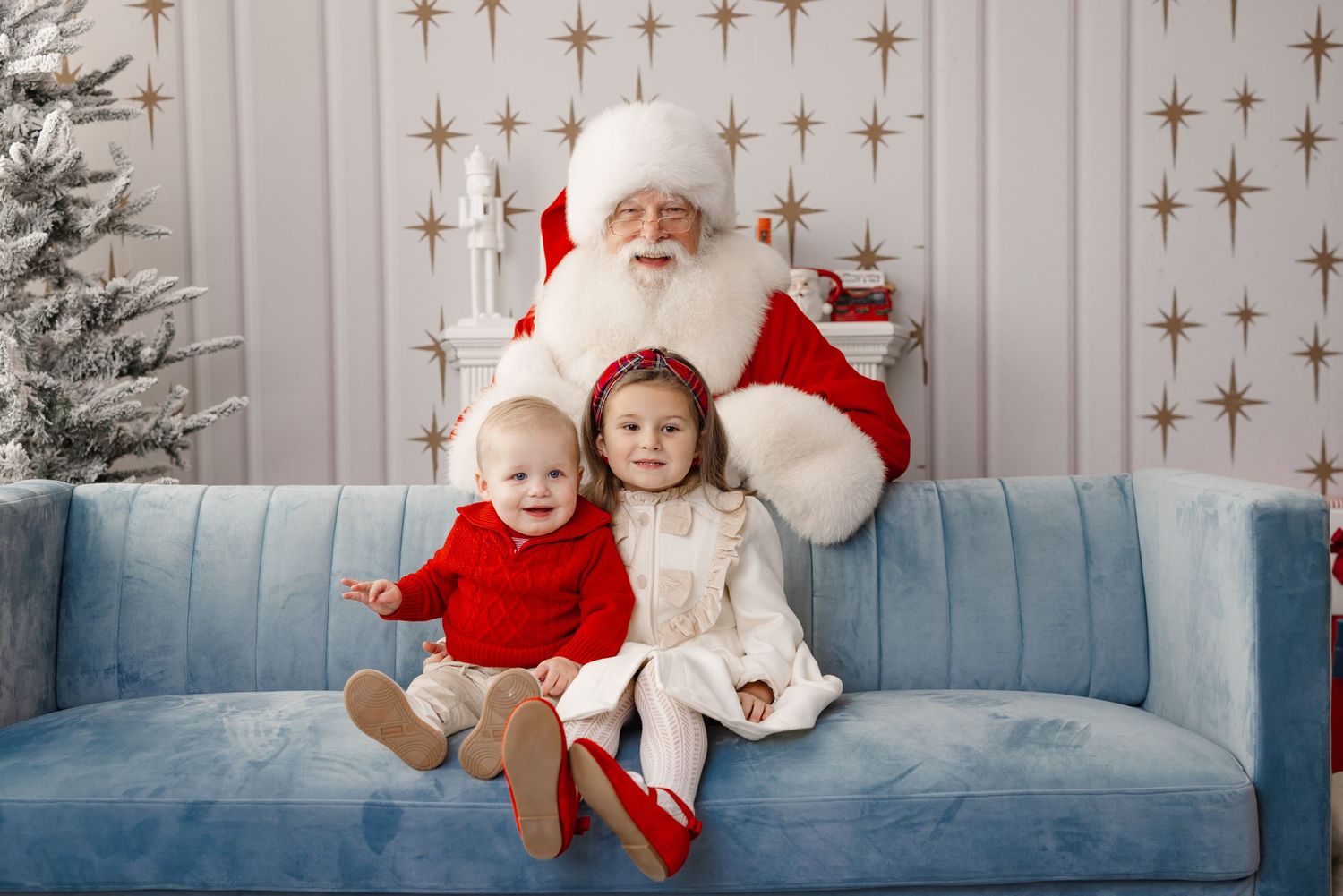 Two children in festive holiday attire sit with Santa Claus on a blue couch against a starry backdrop.